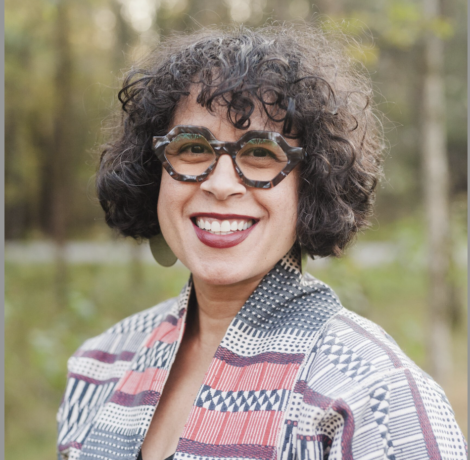 Woman with curly hair and glasses standing for a headshot in front of trees.