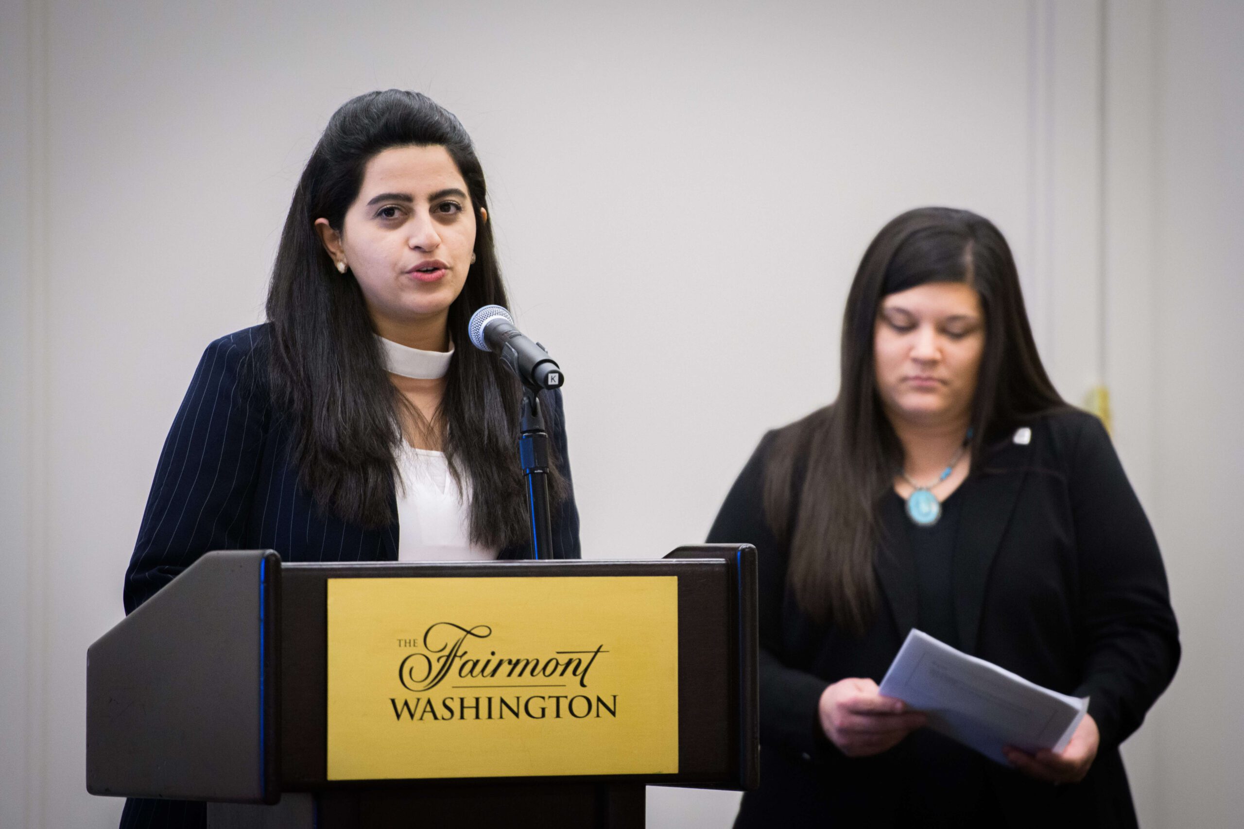 Woman standing in front of a podium speaking into a microphone while another woman looks on.