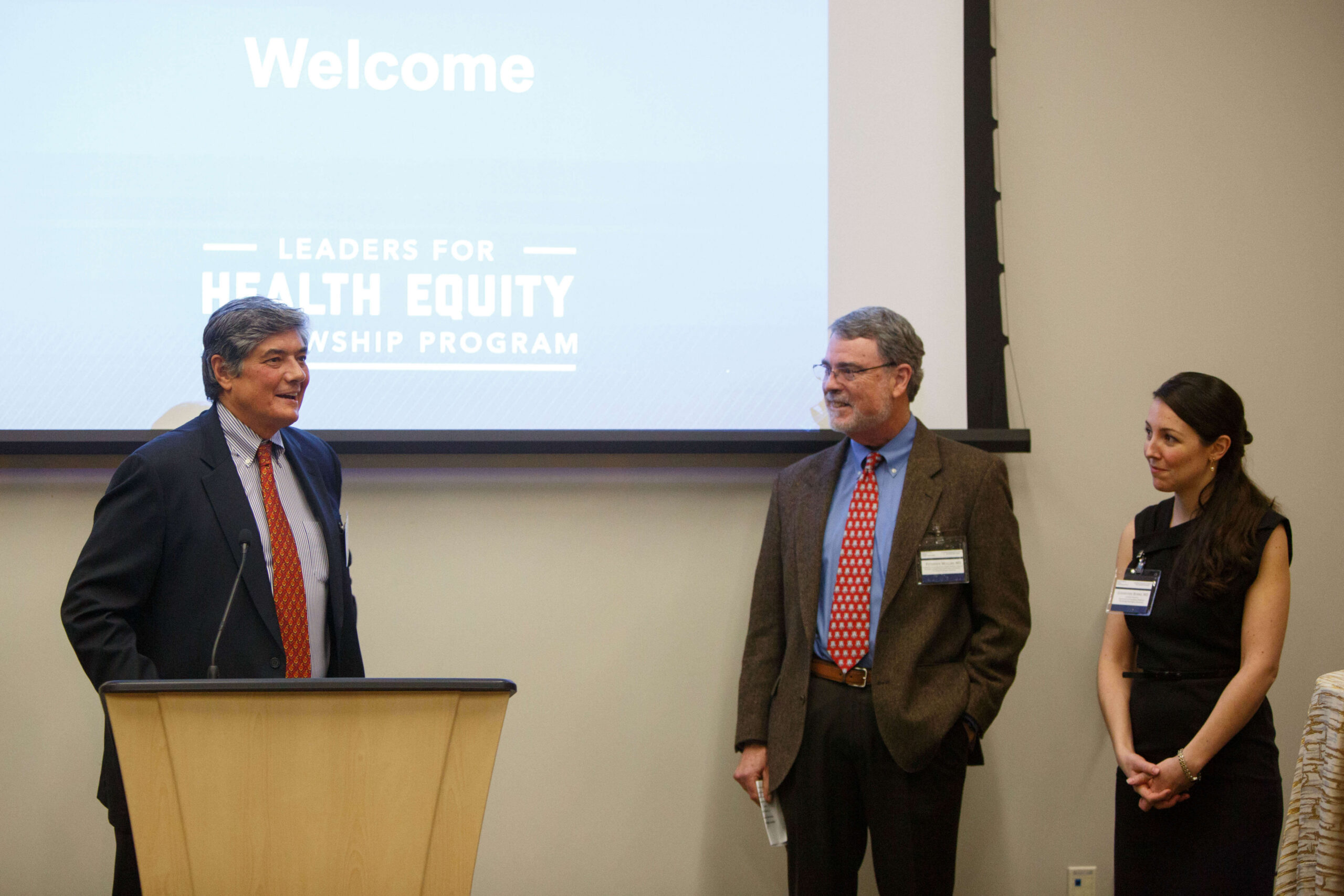 A speaker stands at a podium in front of a screen displaying "Leaders for Health Equity Fellowship Program" while two colleagues stand nearby listening.
