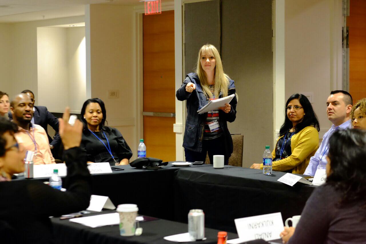 A presenter holding papers points across a conference table toward an attendee raising their hand during a meeting.
