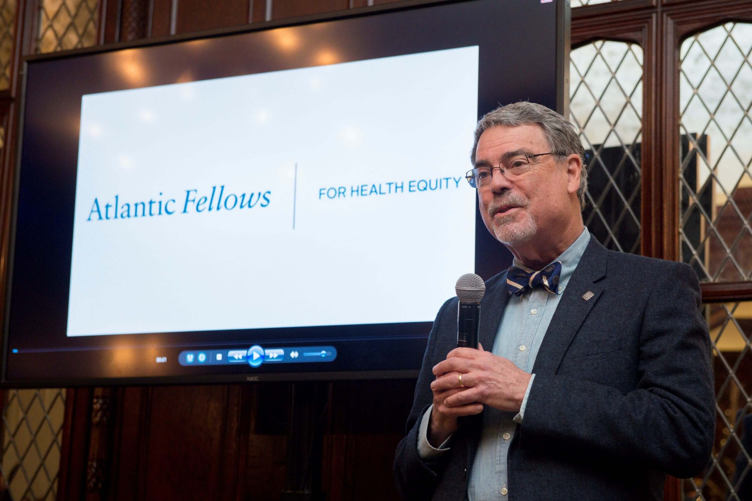 A man in a bow tie speaks into a microphone in front of a screen displaying the Atlantic Fellows for Health Equity logo.