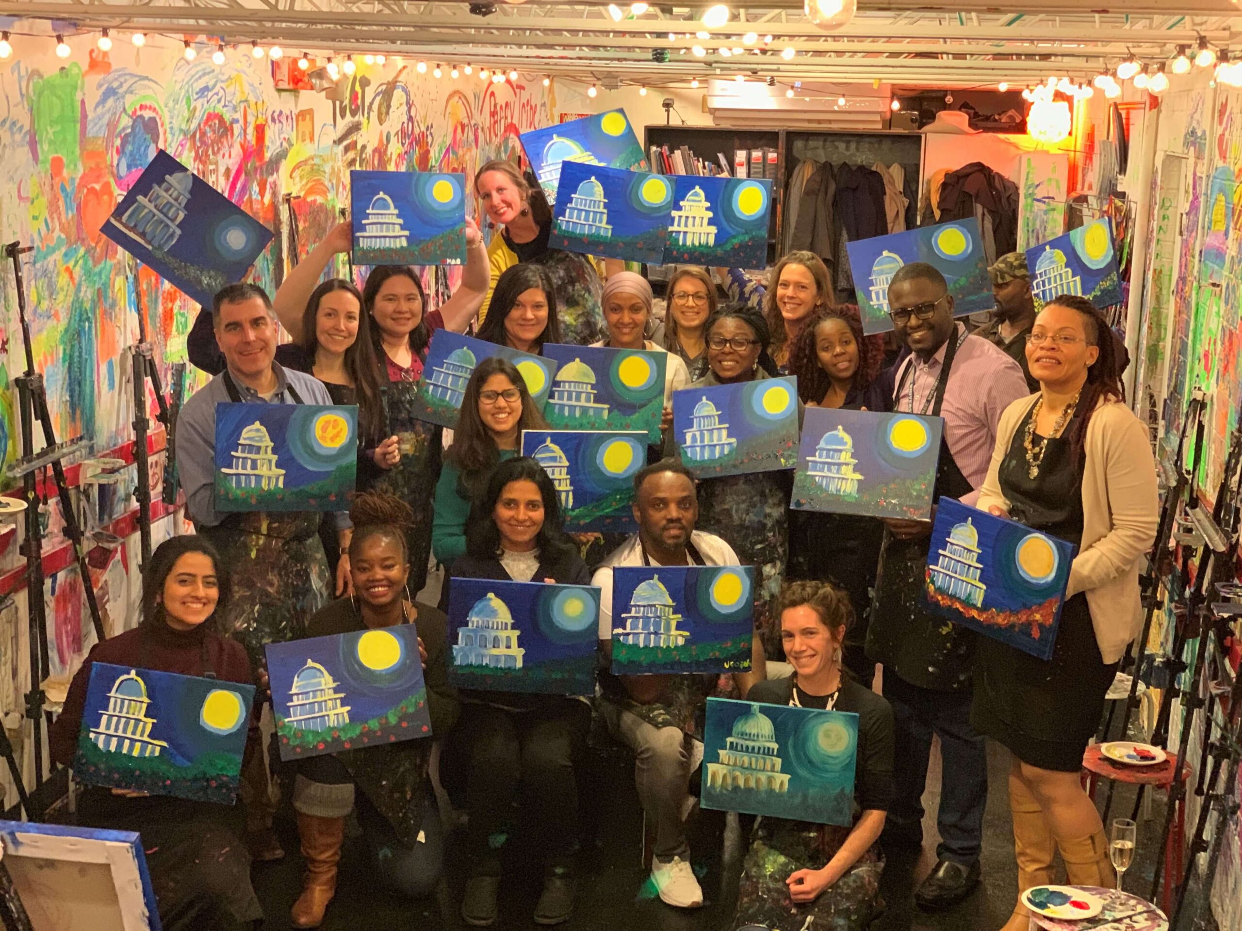 A group of people gather in a brightly lit art studio, smiling and holding up their matching paintings of the U.S. Capitol building at night.