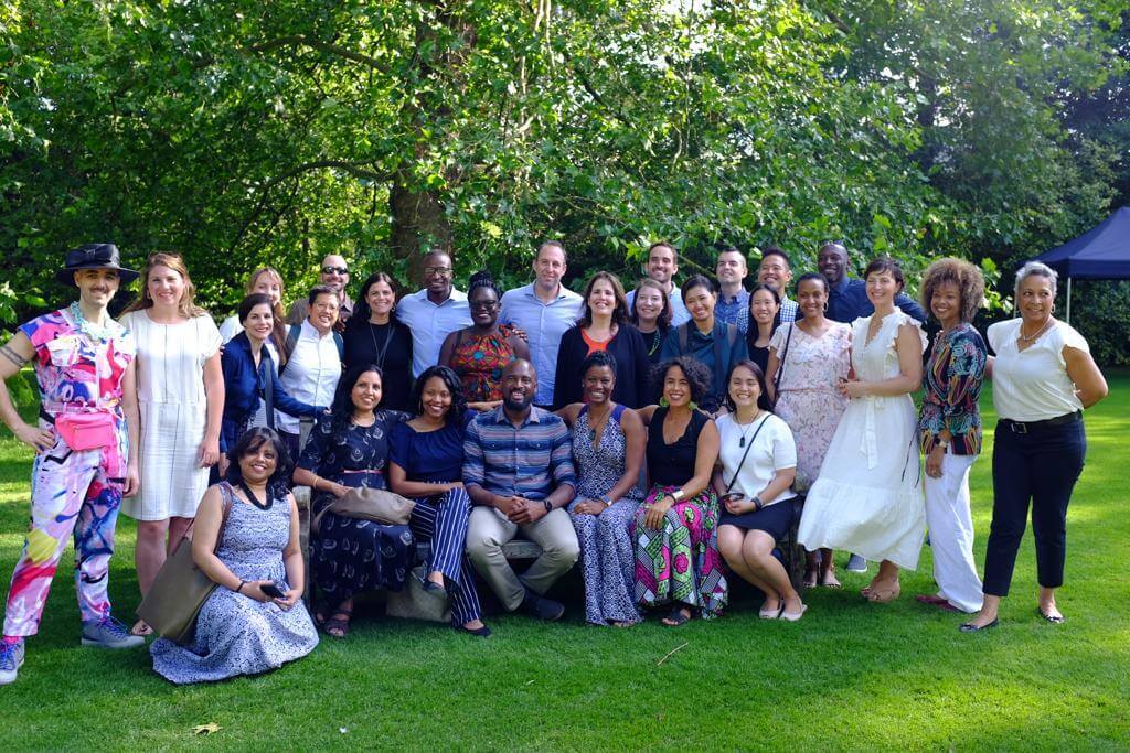 A large group of people pose together on a vibrant green lawn surrounded by lush trees during an outdoor gathering.