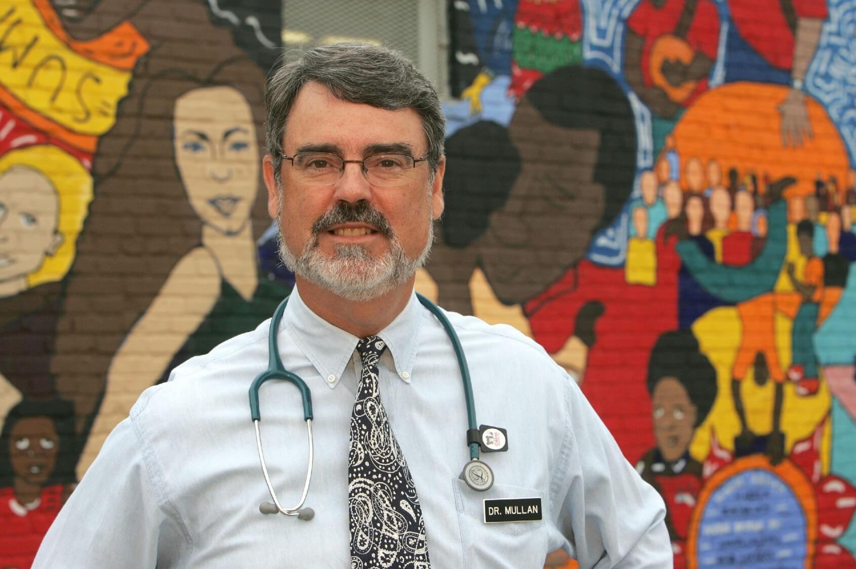 A doctor with a stethoscope around his neck poses in front of a colorful wall mural featuring illustrated figures.