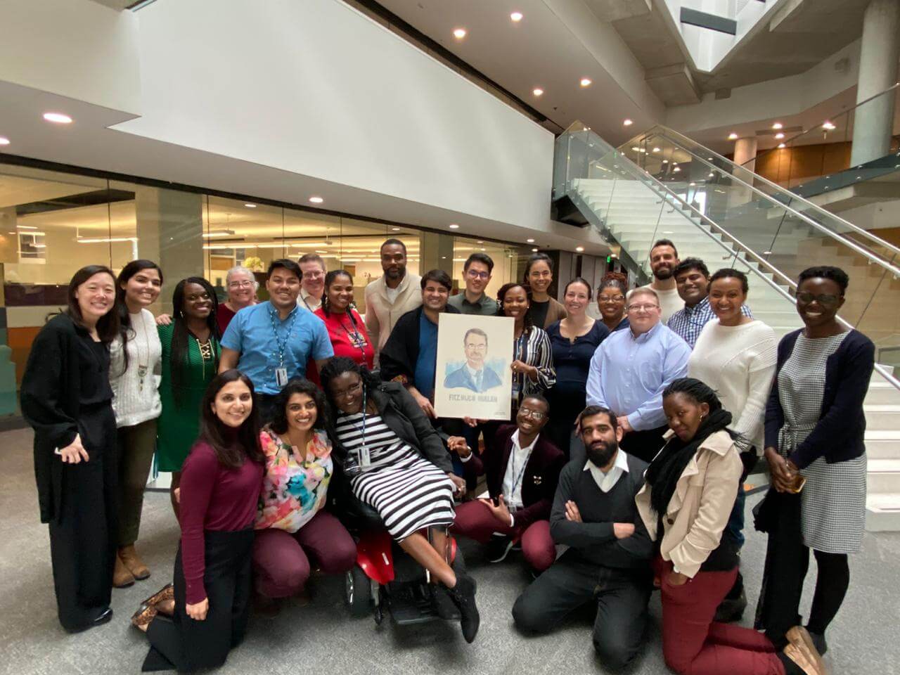 A group of colleagues pose together in a modern building atrium while two members in the center hold up a framed portrait illustration.