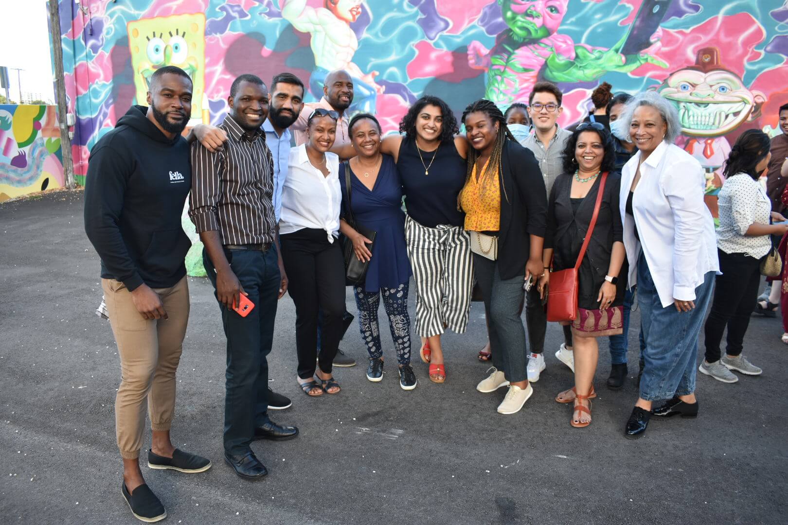 A group of people pose together outdoors on an asphalt surface in front of a colorful, vibrant graffiti wall.