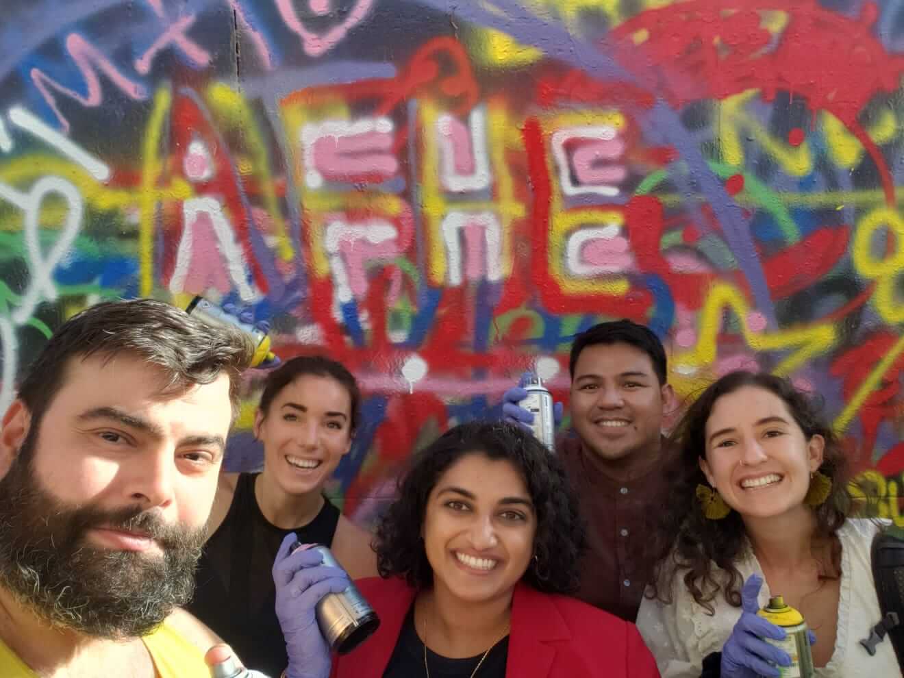 Five smiling individuals pose for a selfie while holding spray paint cans in front of a colorful, graffiti-covered wall.