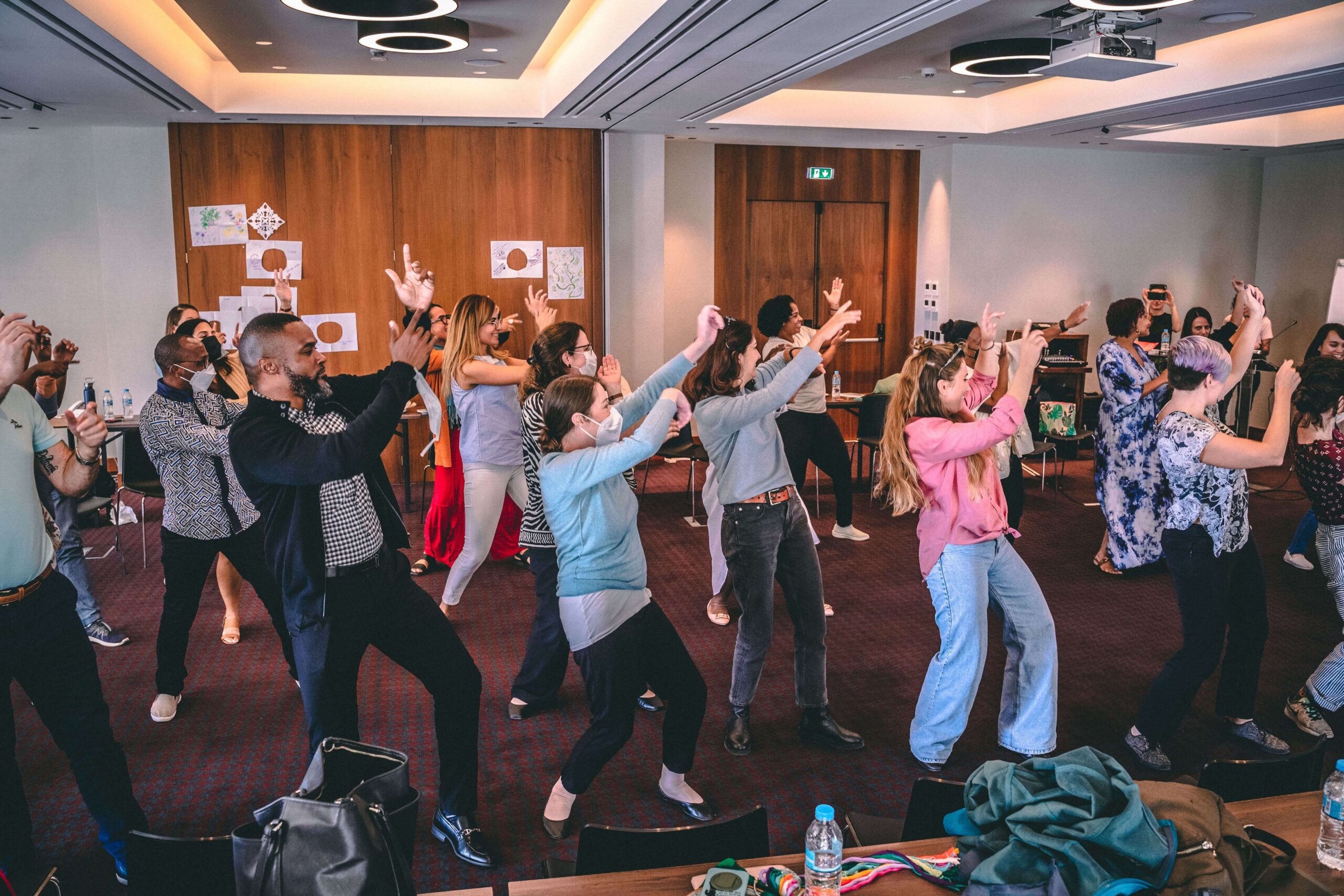 A group of people practice synchronized dance moves inside a spacious conference room with dark red carpeting.