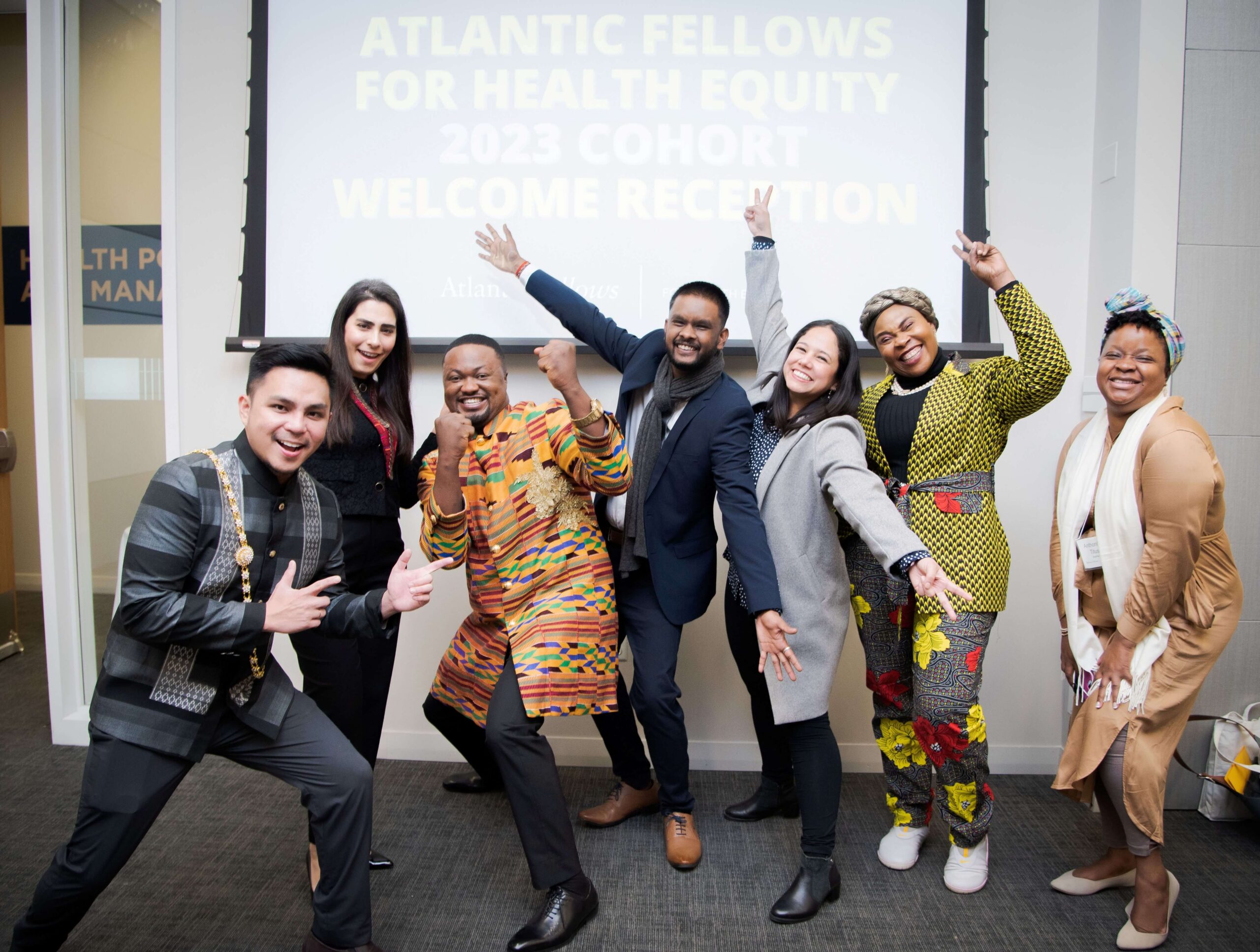 Seven group members pose joyfully in front of a projection screen announcing the Atlantic Fellows for Health Equity 2023 Cohort Welcome Reception.