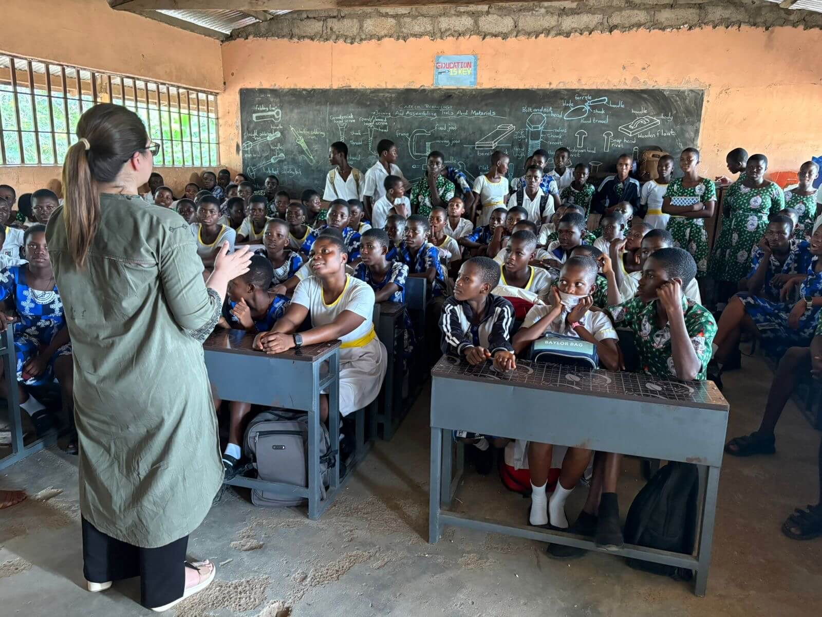 A presenter stands at the front of a crowded classroom addressing dozens of students seated at wooden desks.