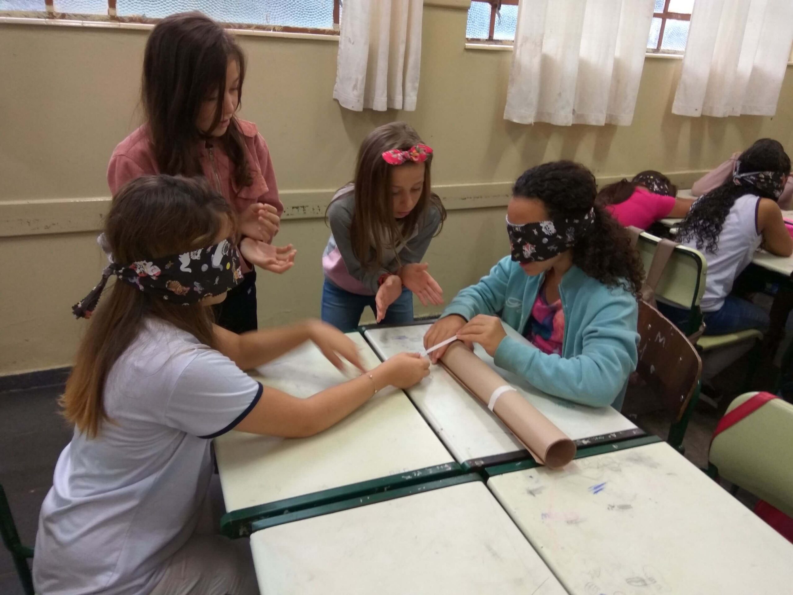 Four young girls in a classroom participating in a sensory activity, with two blindfolded girls at desks working together to tie a string around a rolled-up paper while two classmates watch and guide them.