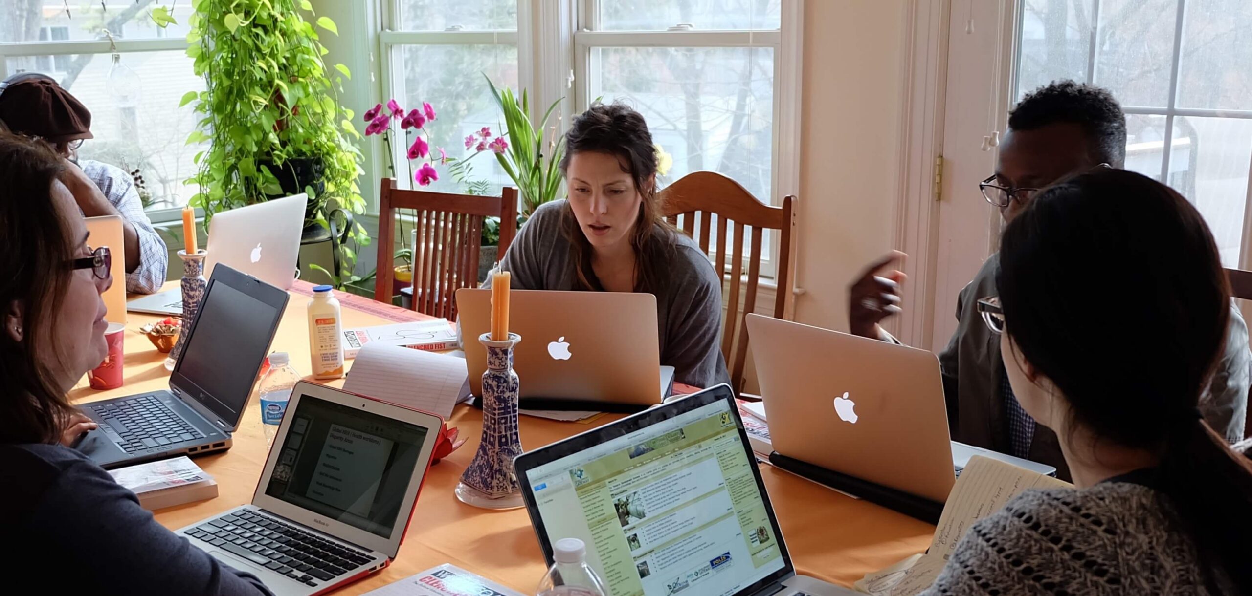 A group of diverse people sitting around a large dining table filled with laptops, notebooks, and plants, engaged in a collaborative workspace session indoors
