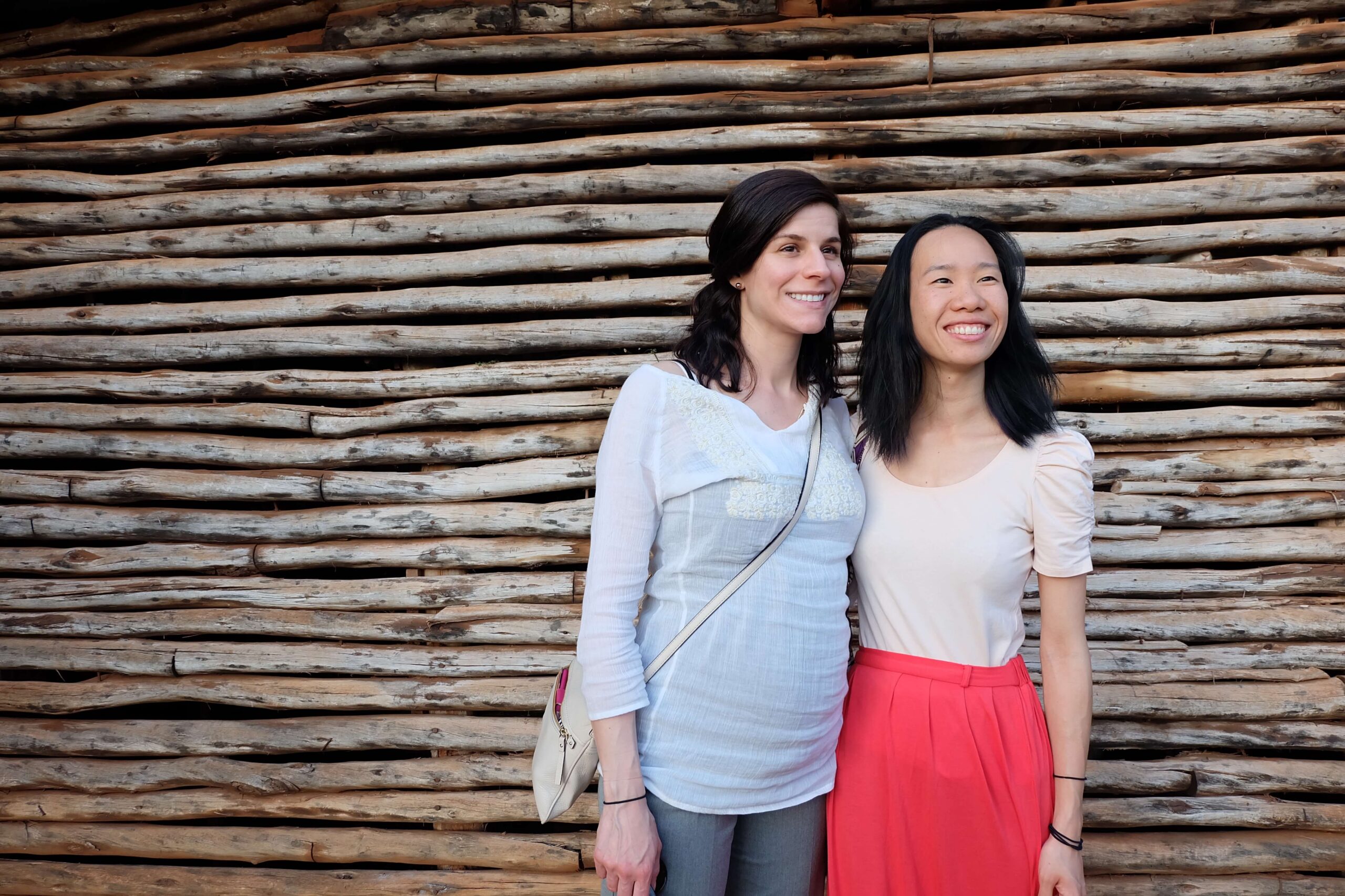 Two women smiling and posing together outdoors in front of a wall made of stacked wooden logs.
