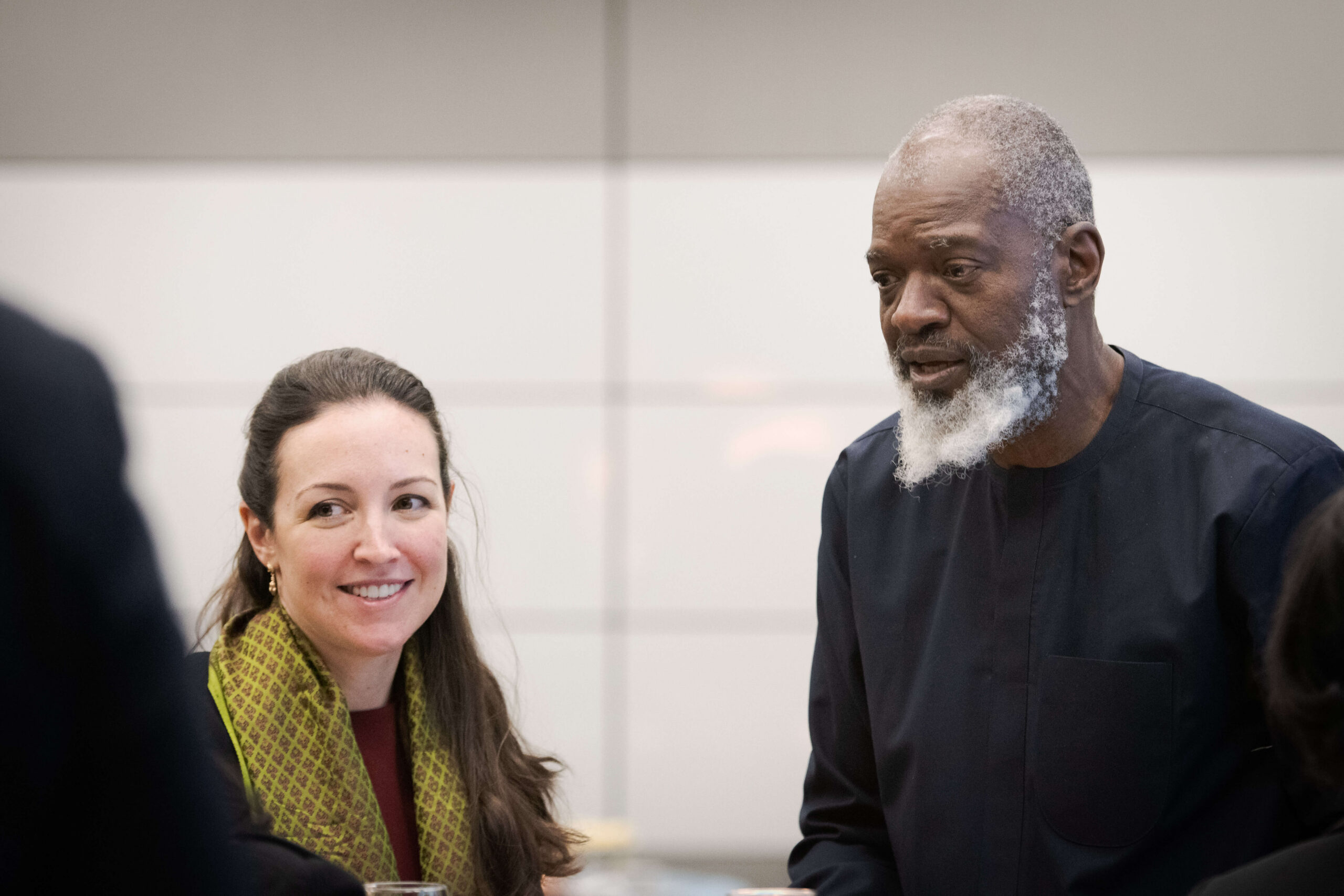 A woman with a patterned green scarf smiles warm-heartedly while looking toward a man with a white beard wearing a dark long-sleeved shirt.