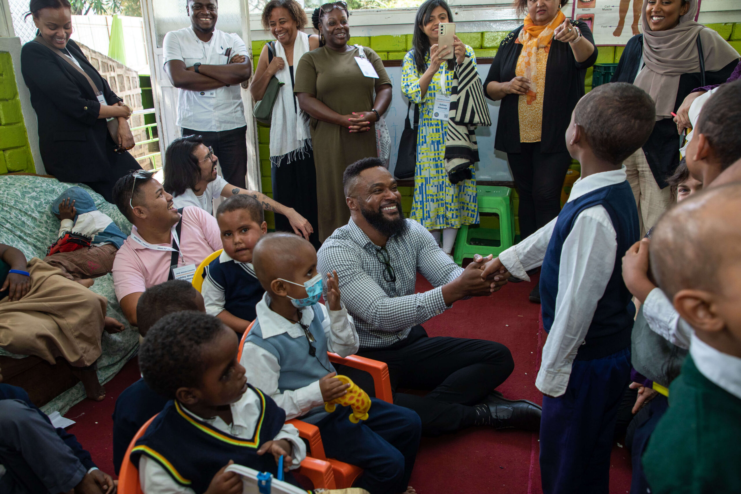 A man sitting on the floor smiling and shaking hands with a young boy surrounded by children and adult observers indoors.