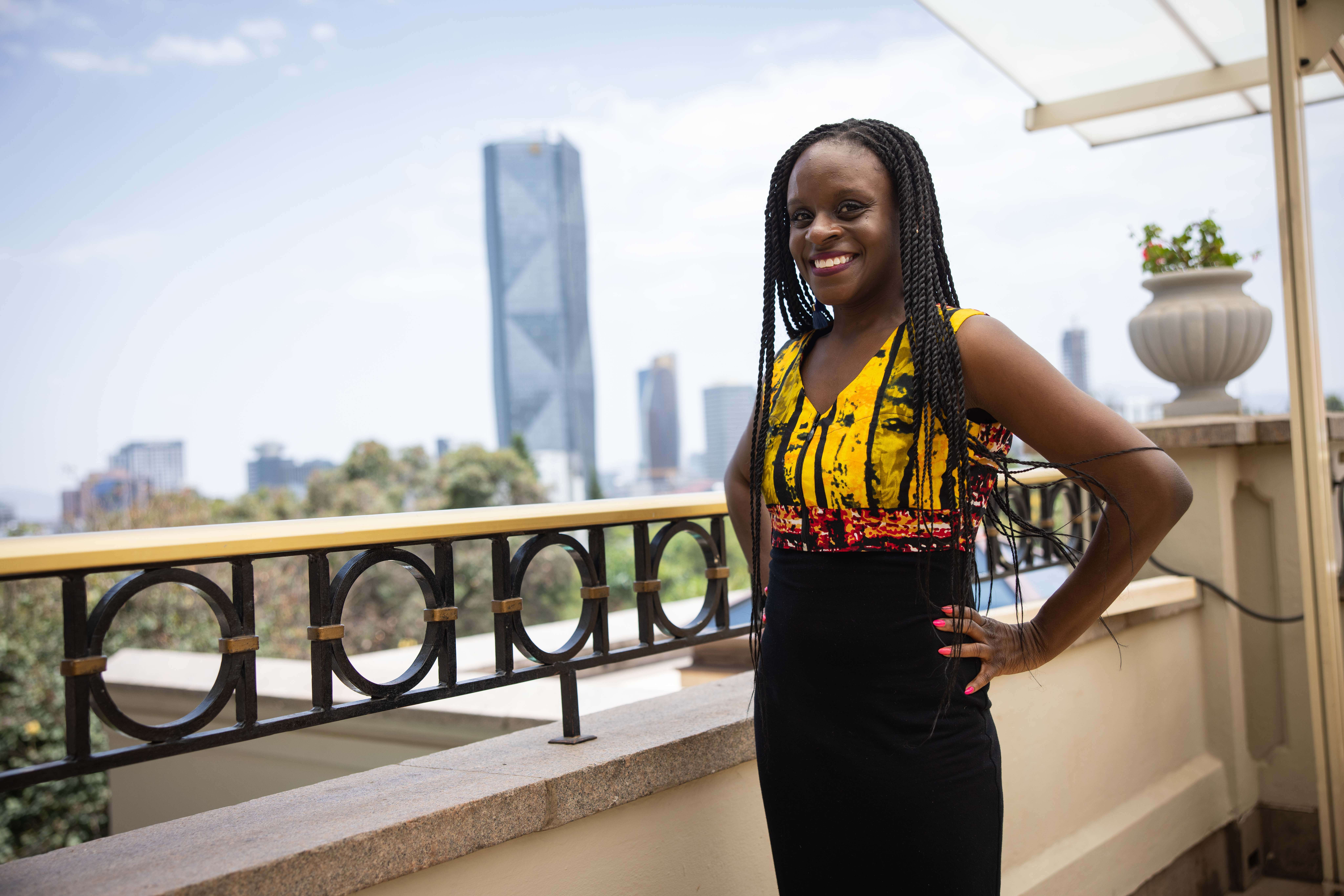 A smiling woman with long braided hair posing on a balcony overlooking a city skyline.