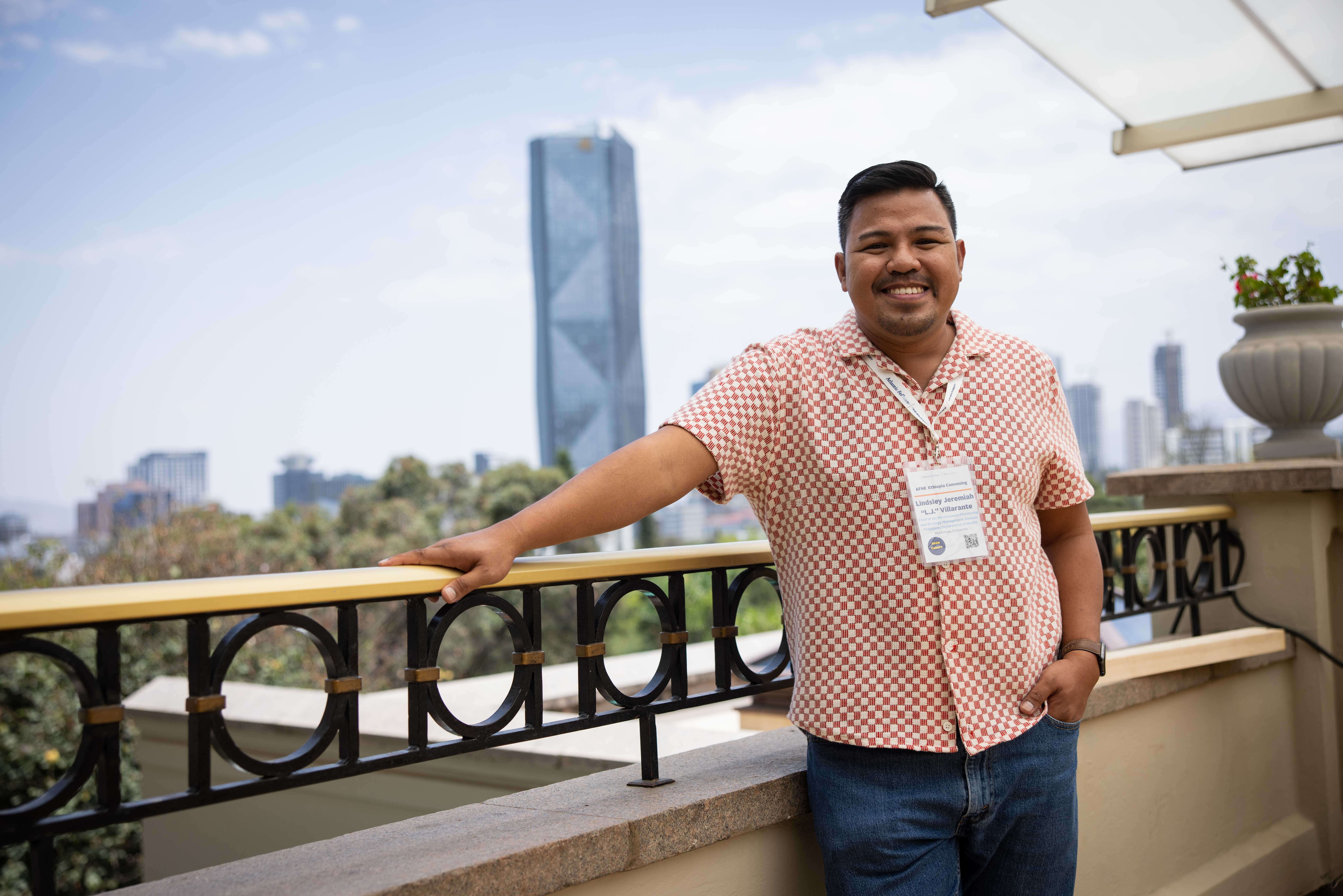 A smiling man wearing a patterned shirt and lanyard leaning against a balcony railing with a city skyline behind him.