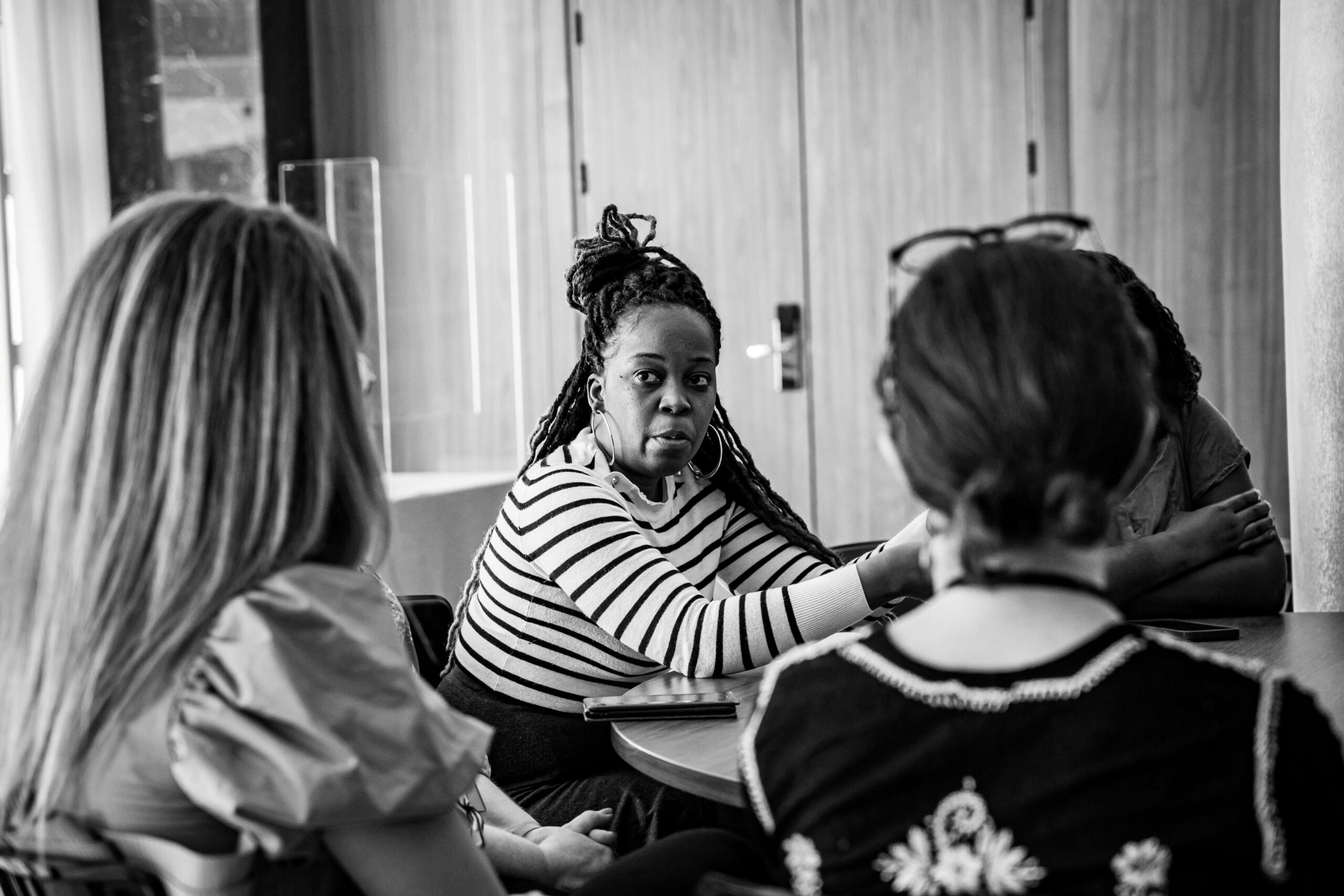 Black-and-white photo of a small group seated at a table in discussion, focusing on a woman with dreadlocks in a striped shirt speaking.