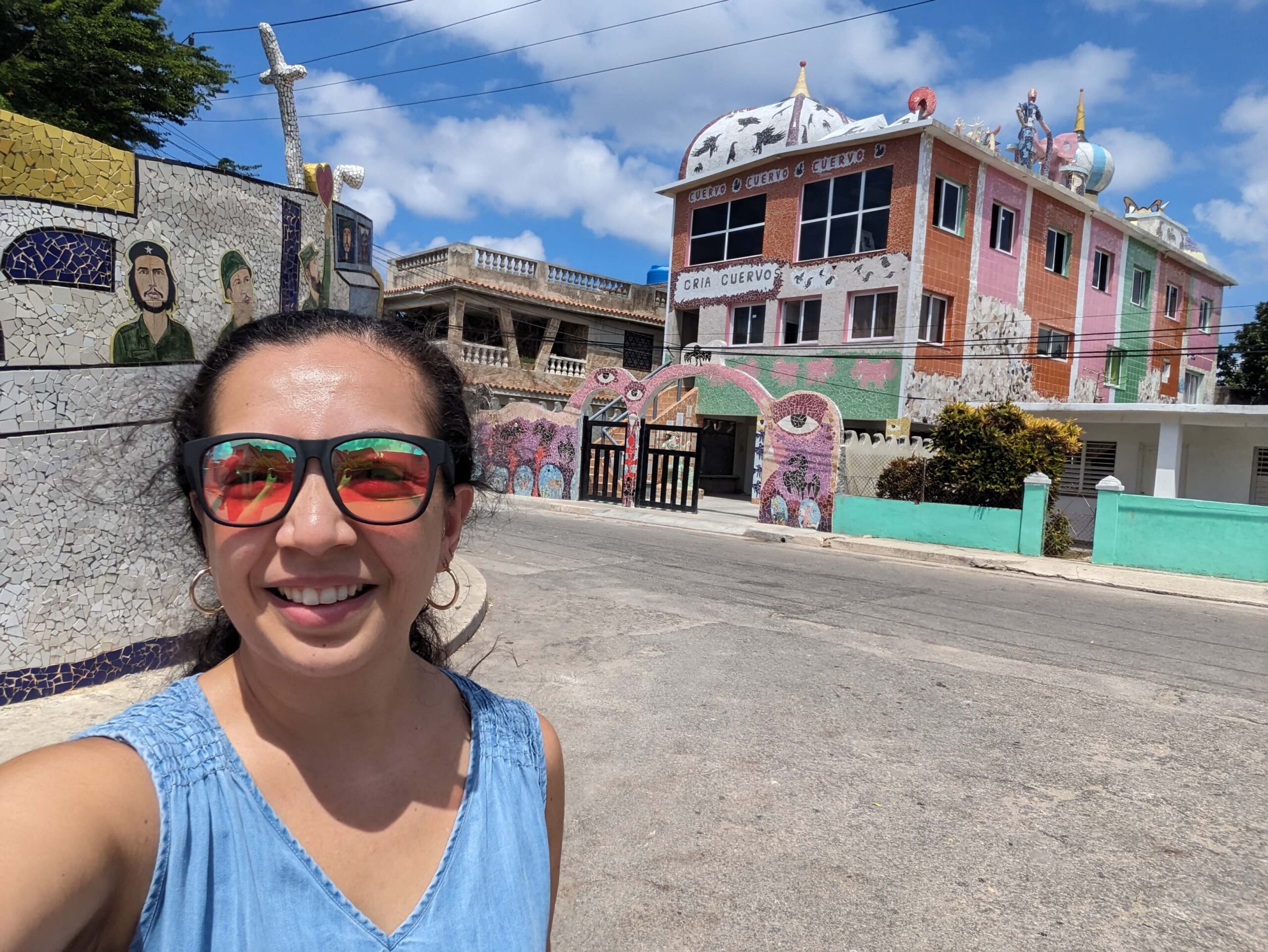 A smiling woman in a blue tank top and reflective sunglasses taking a selfie on a street with vibrant, mosaic-tiled colorful buildings (Fusterlandia in Havana, Cuba).