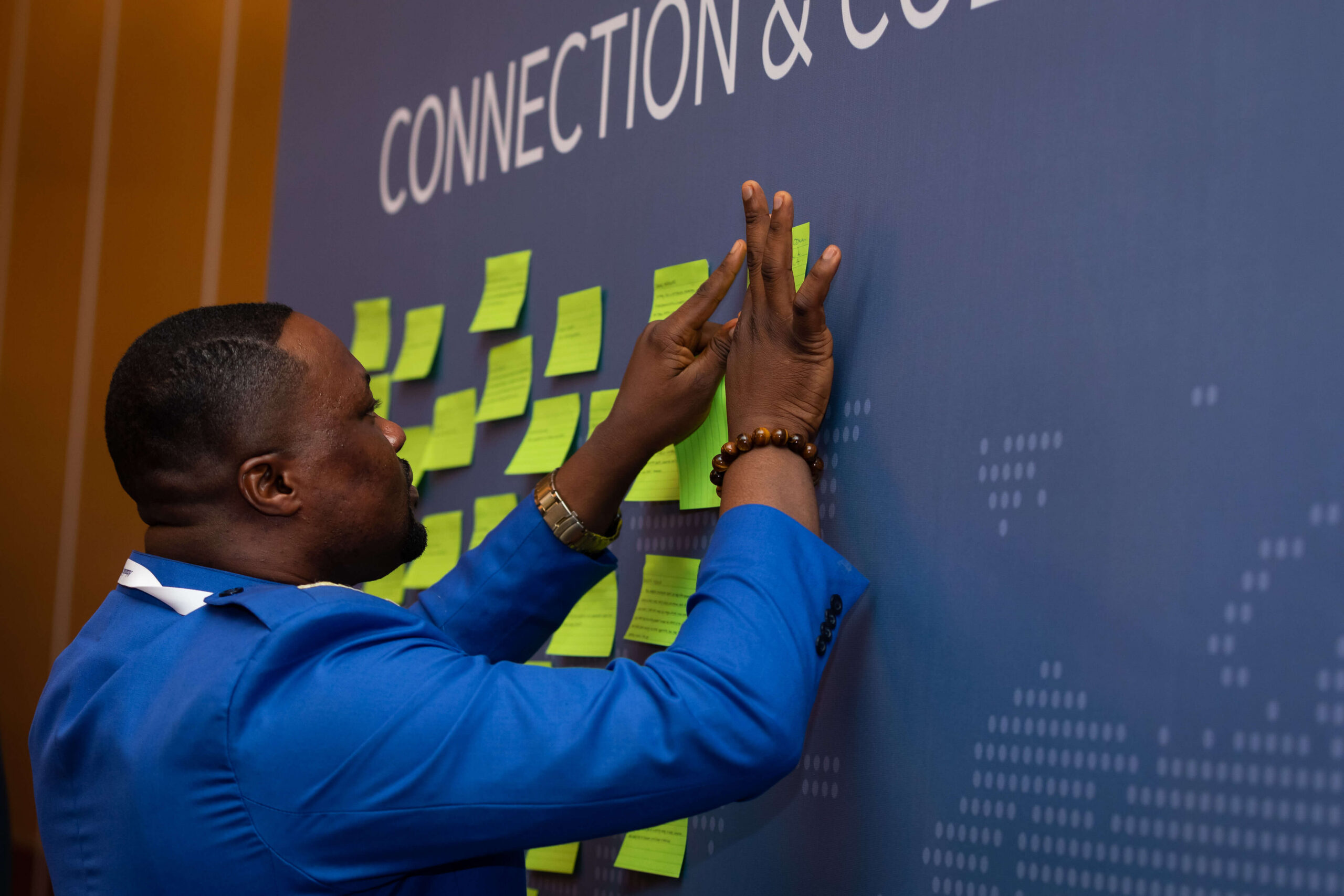 Man in a bright blue jacket placing green sticky notes on a presentation wall displaying the word 