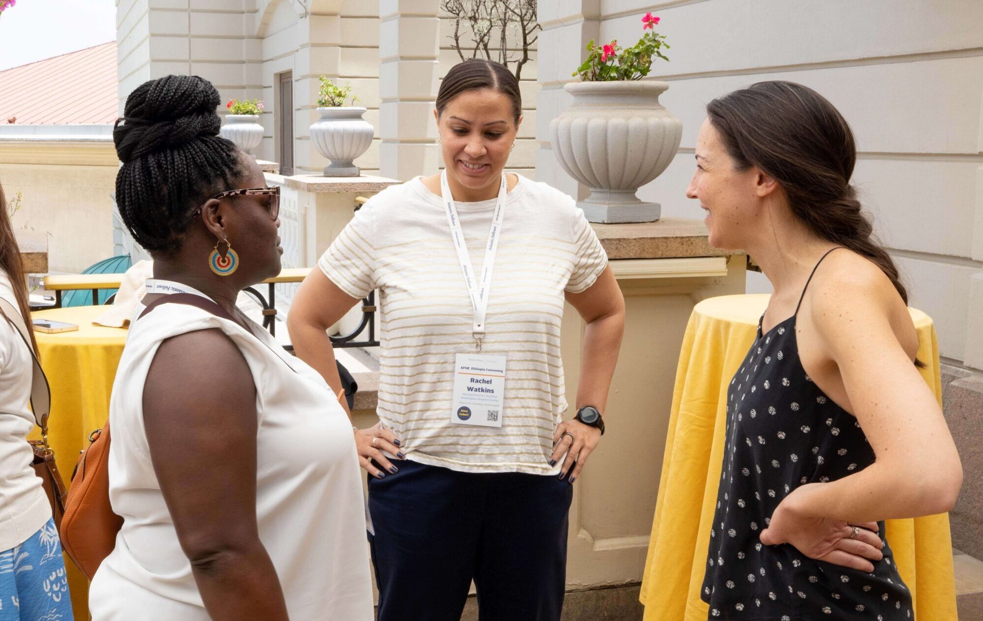 Three women stand talking outdoors near yellow-clothed tables and potted plants.
