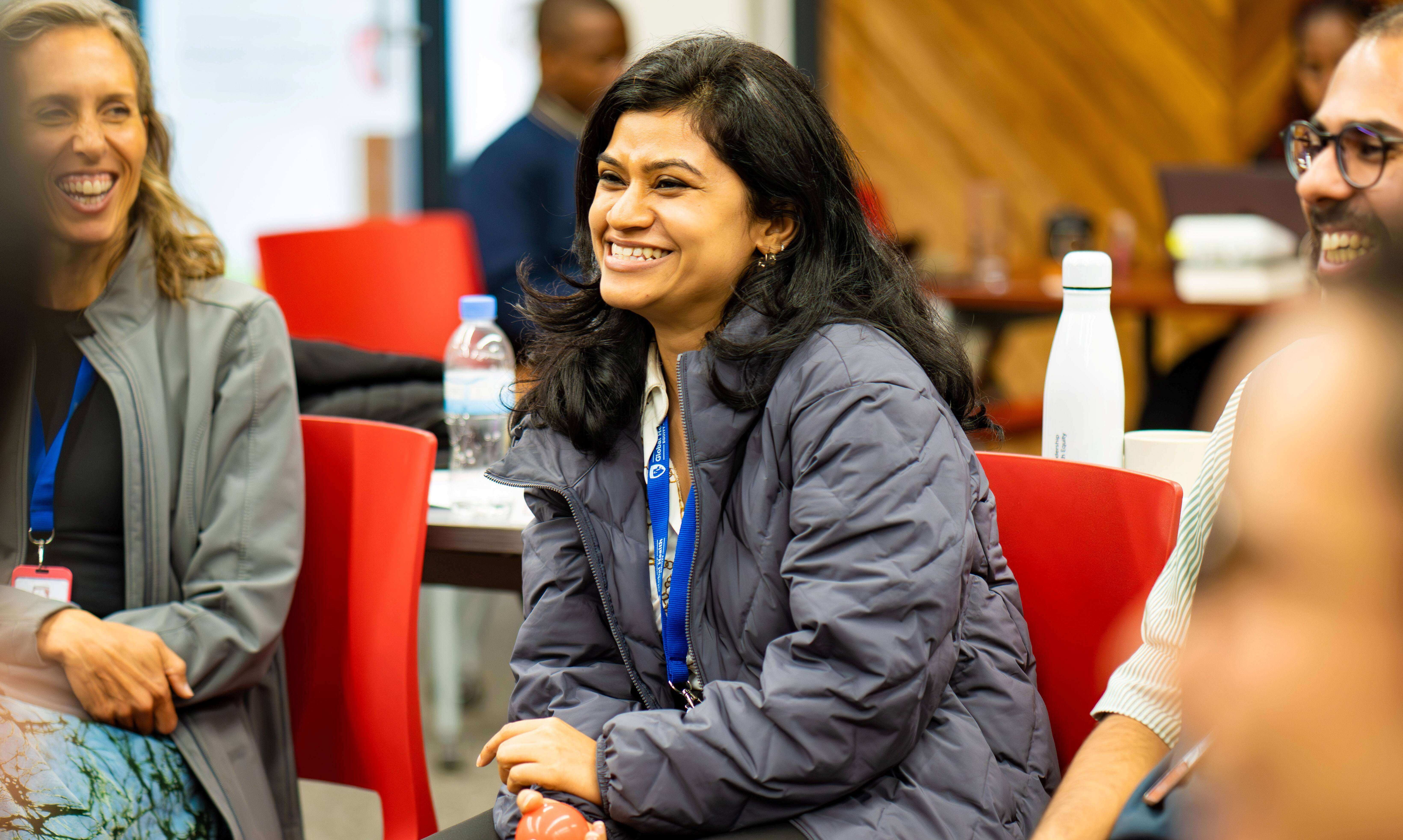 A smiling woman with dark shoulder-length hair wearing a grey puffer jacket, seated in a group circle and holding a small orange ball during a workshop session.