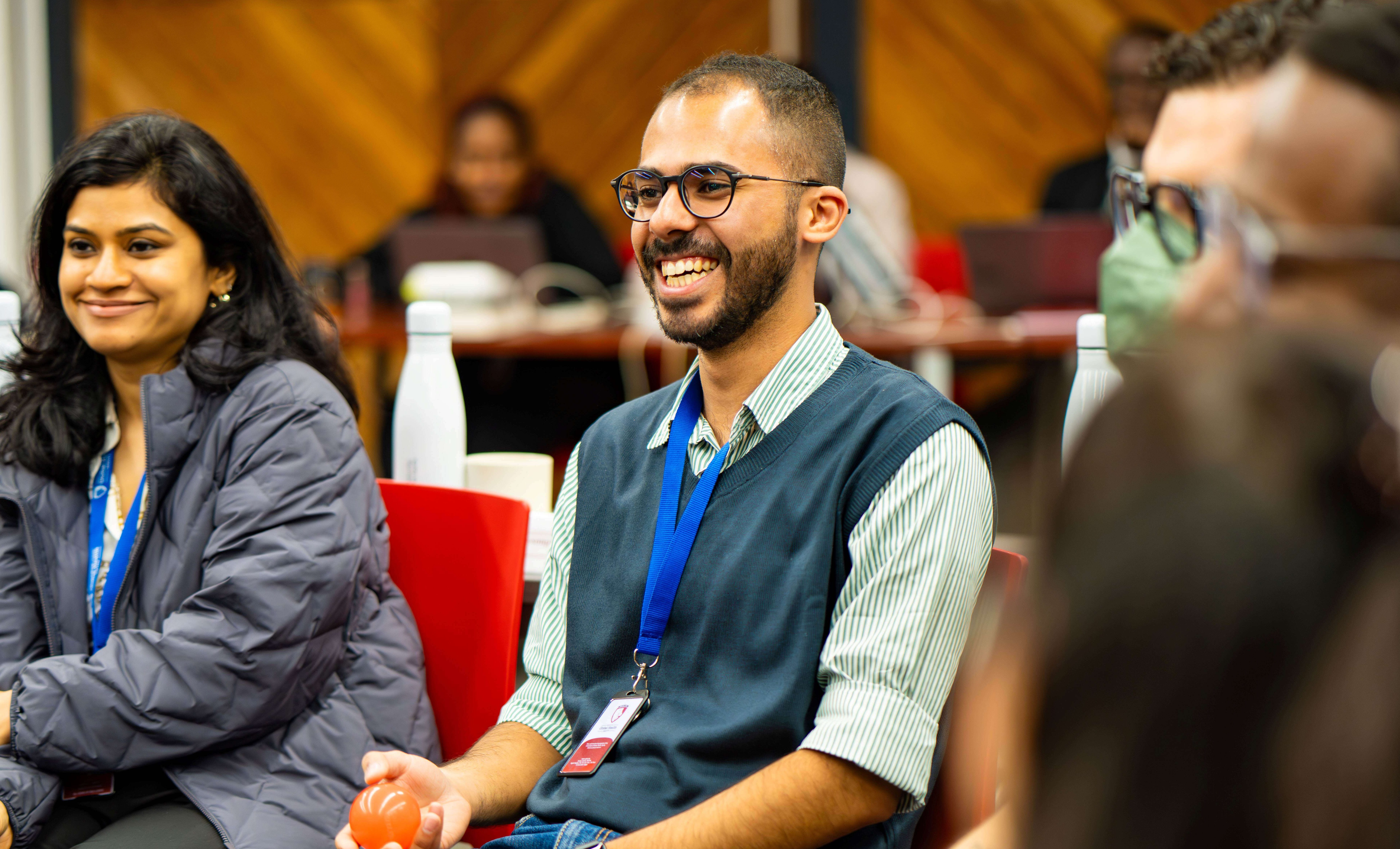 A smiling man in a blue lanyard holding a small orange ball while speaking to fellow participants seated in red chairs.
