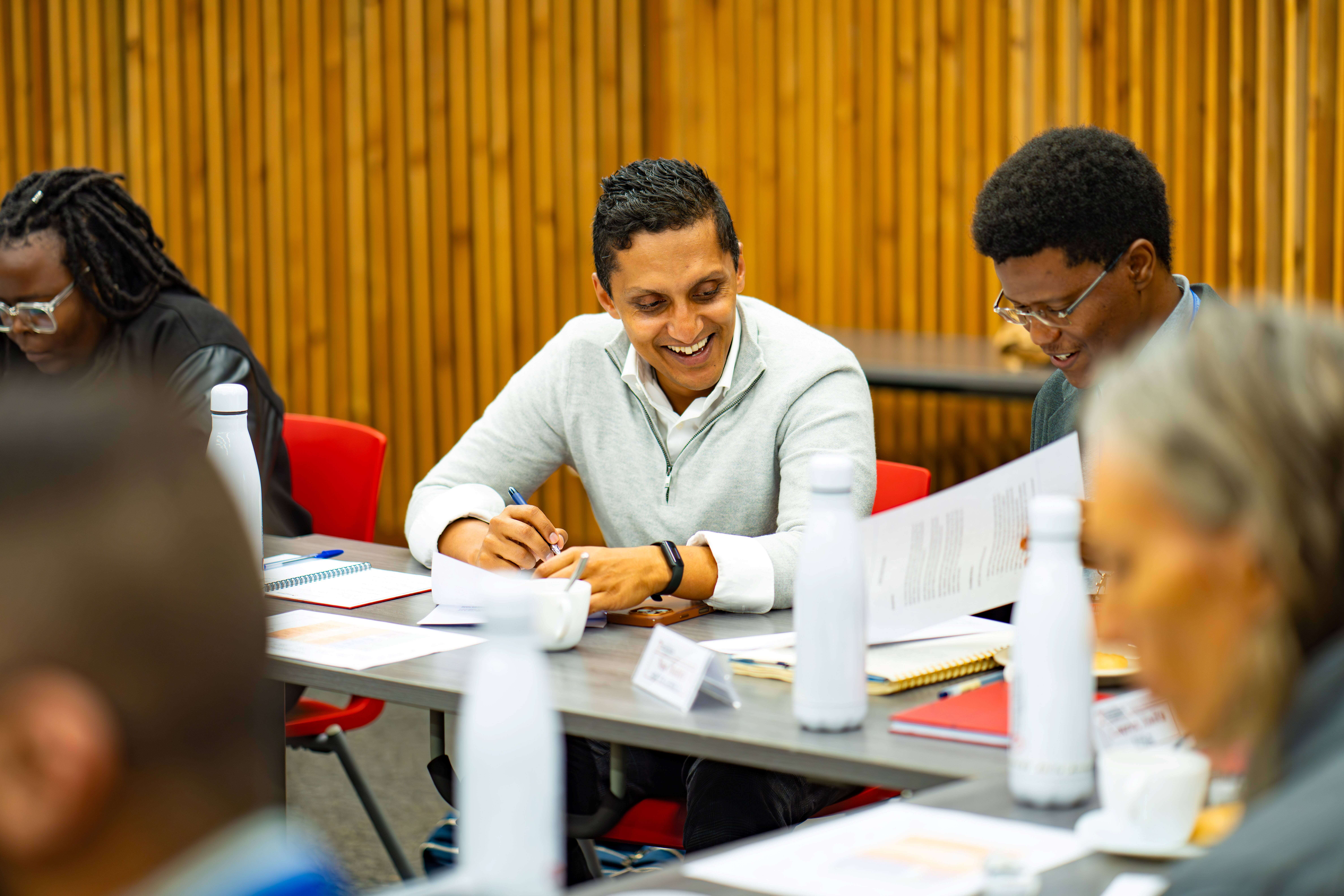 A smiling man in a light grey quarter-zip sweater sitting at a conference table writing in a notebook alongside other meeting attendees.