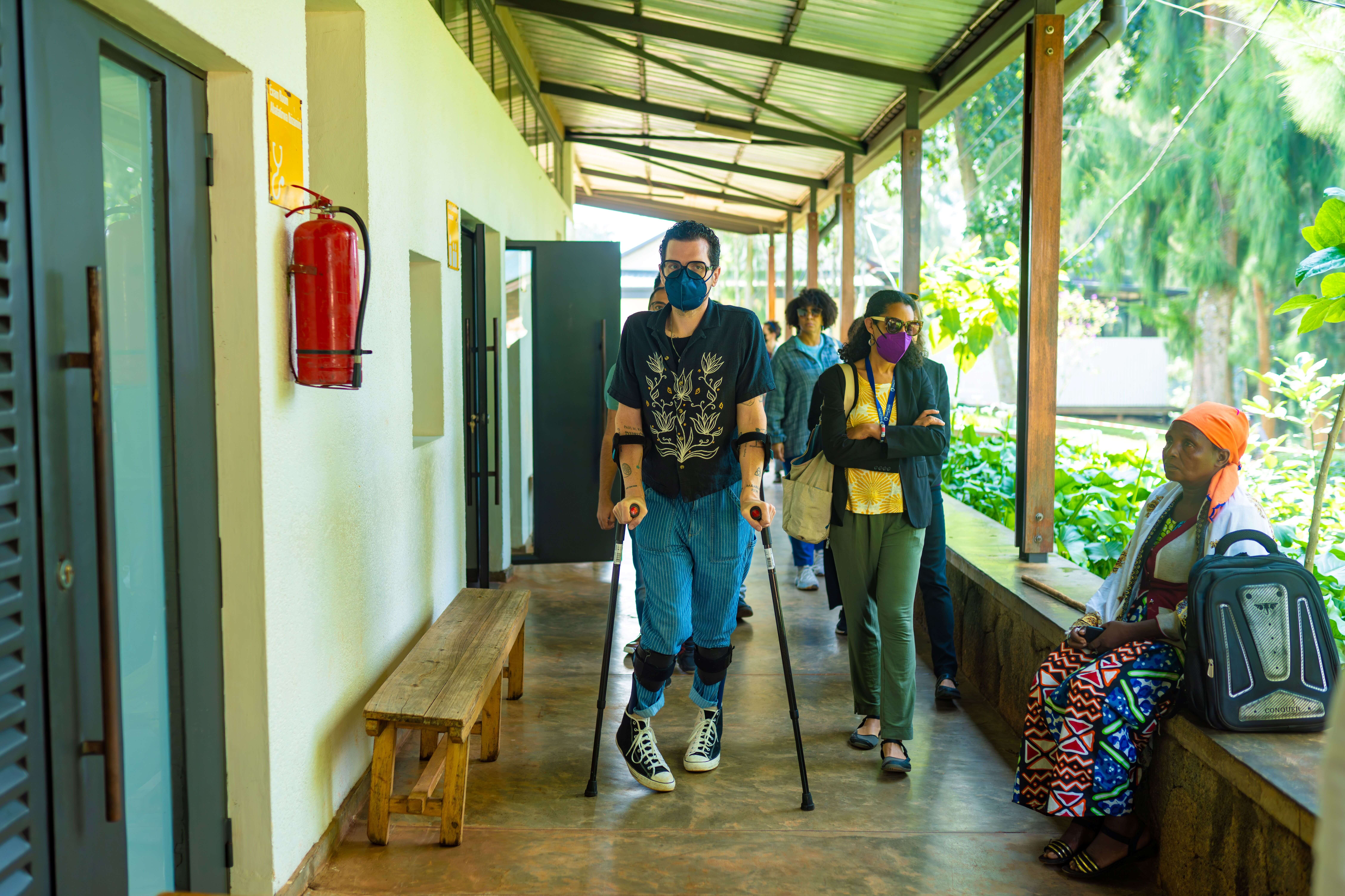 A person wearing a face mask, patterned shirt, blue trousers, and leg braces walking down a covered outdoor walkway using forearm crutches while accompanied by colleagues.