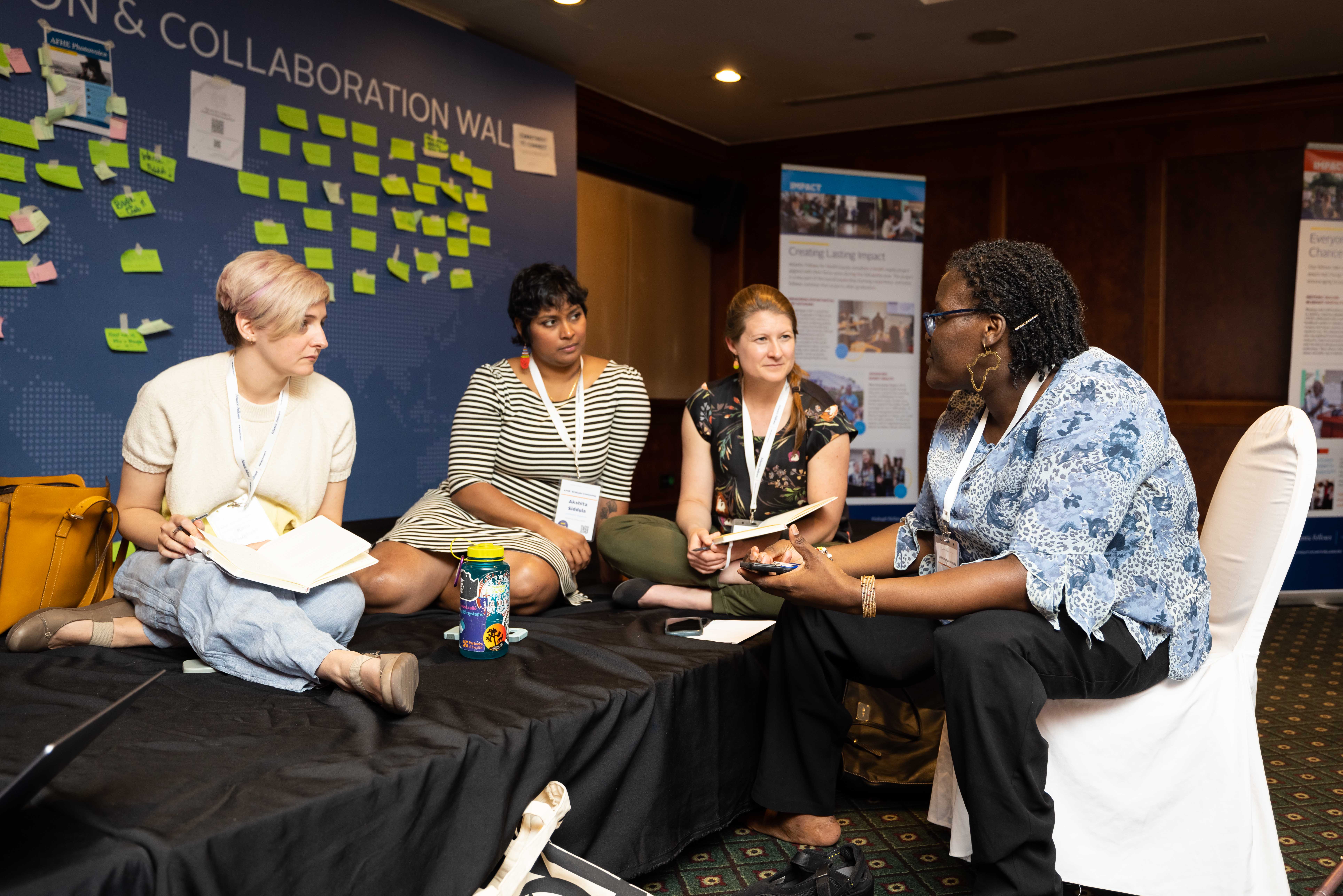 Four people sit together in a group discussion in front of a sticky-note wall.