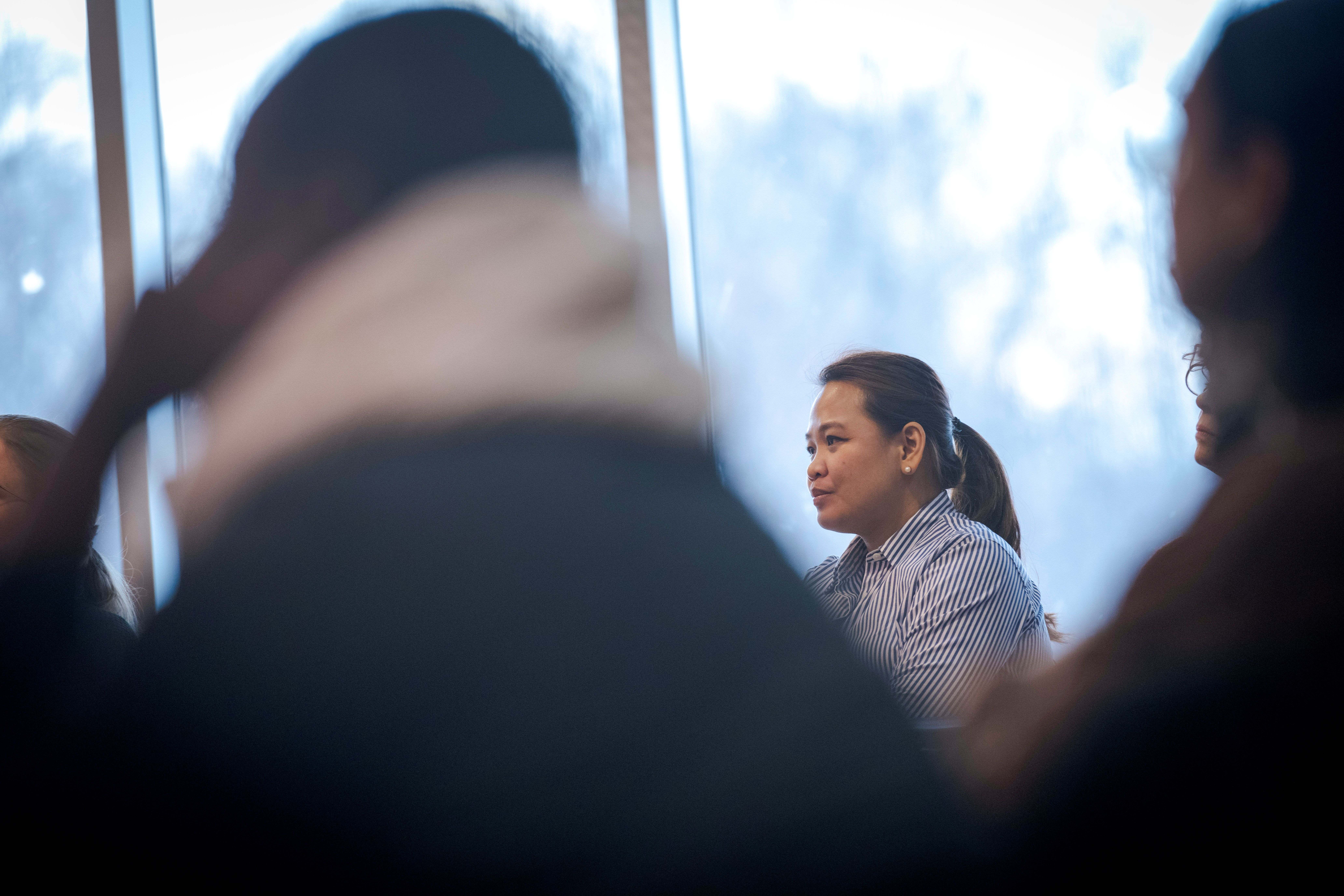 A woman in a striped shirt sits near a window, framed by out-of-focus meeting participants.