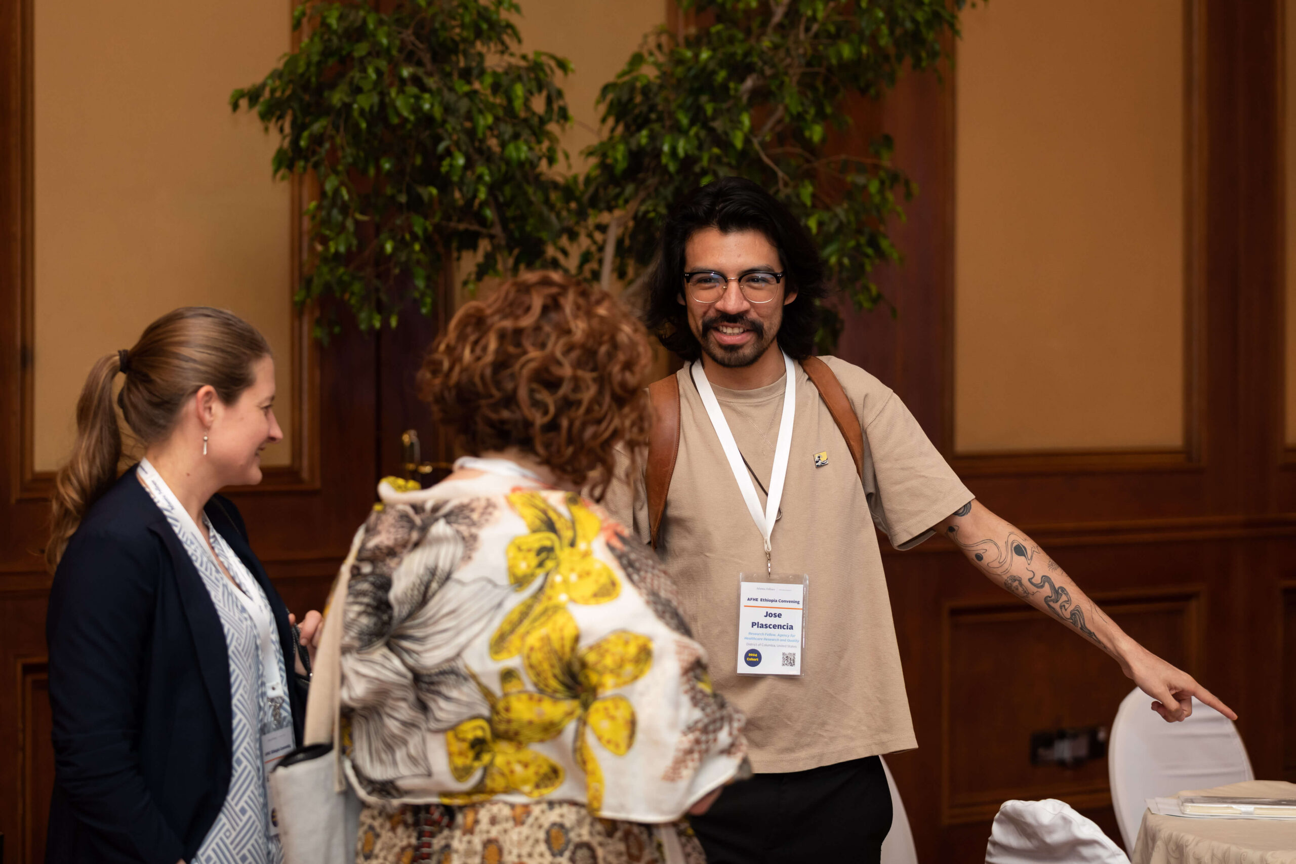 A man in a beige shirt and glasses smiles and points while talking to two women.