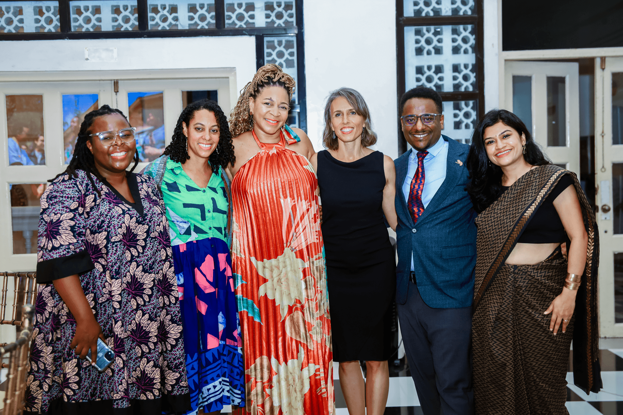 Six smiling colleagues posing together indoors for a group photo, dressed in colorful patterned dresses, formal suits, and a traditional saree