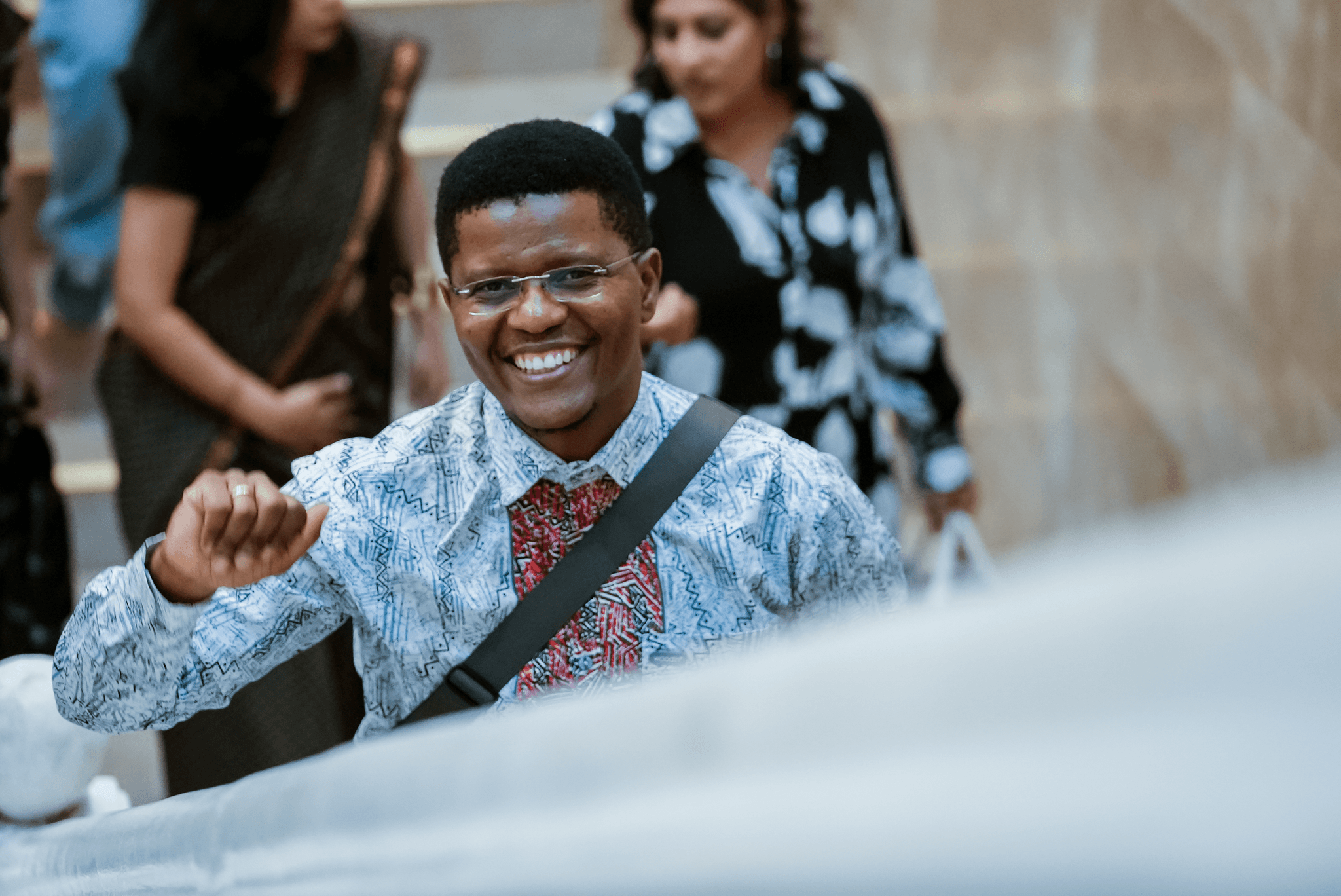 A smiling man with glasses wearing a white patterned shirt with red trim holding up a light fist gesture while walking up a staircase.
