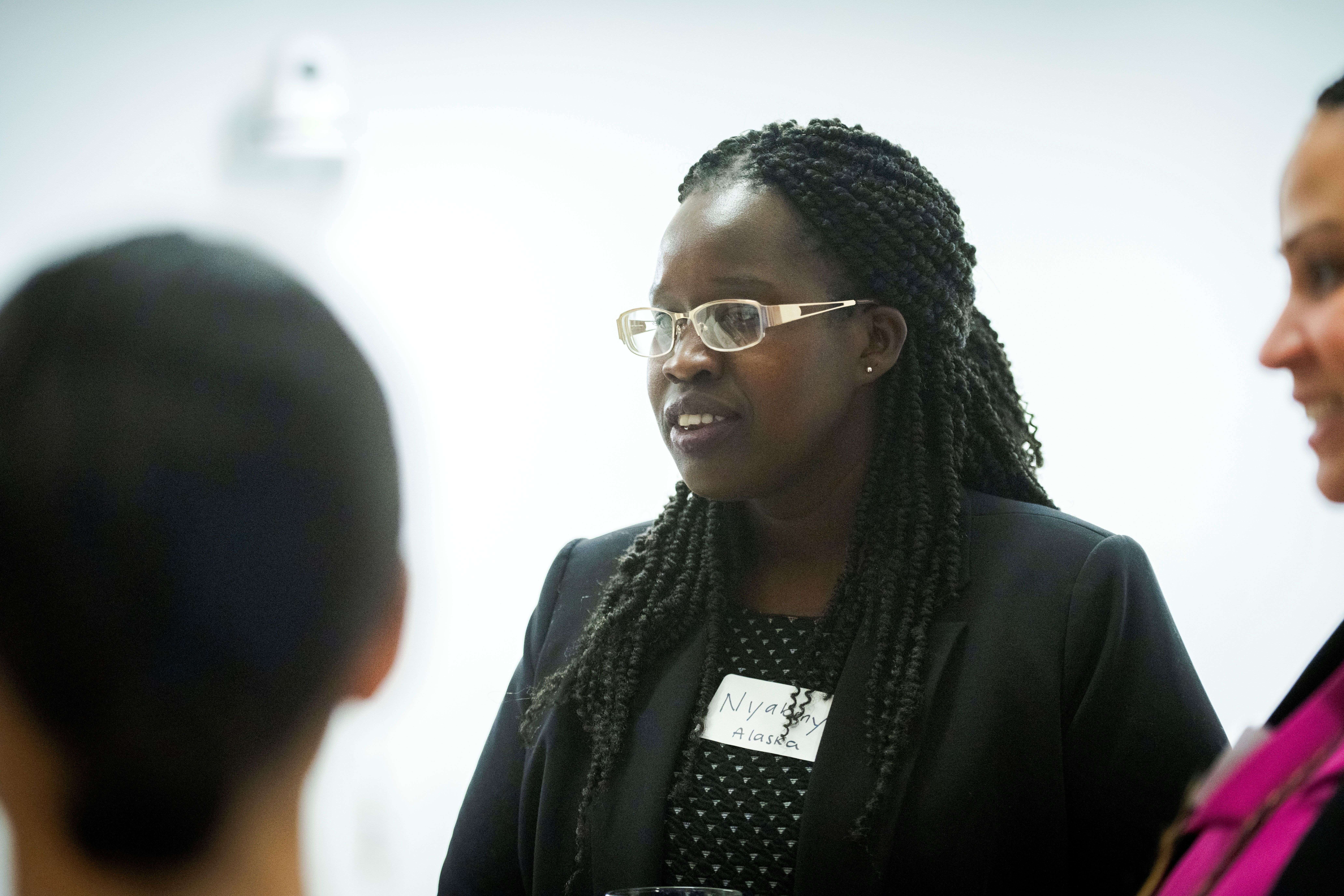 A woman with braided hair and glasses wearing a name tag speaks to nearby colleagues.