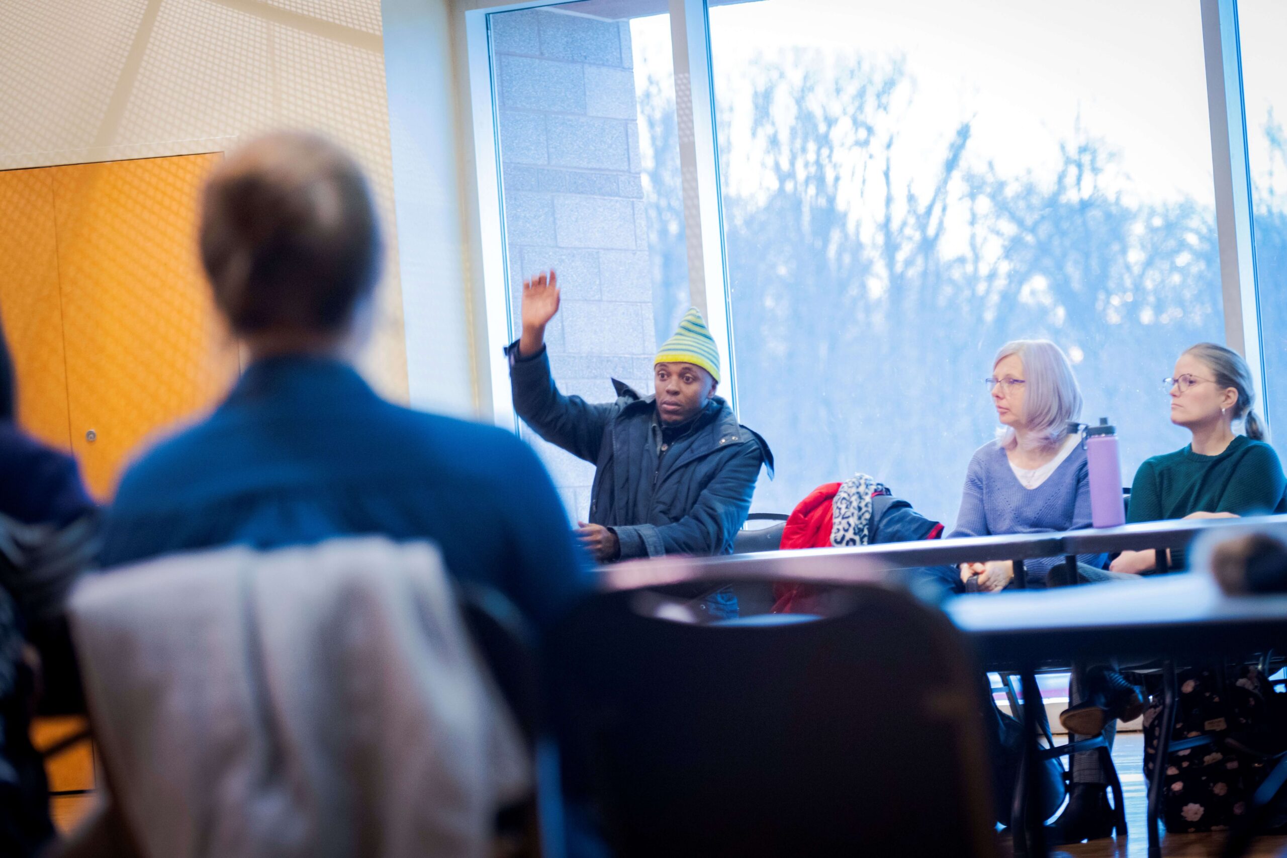 A man in a striped beanie raises his hand while seated at a table during a meeting.