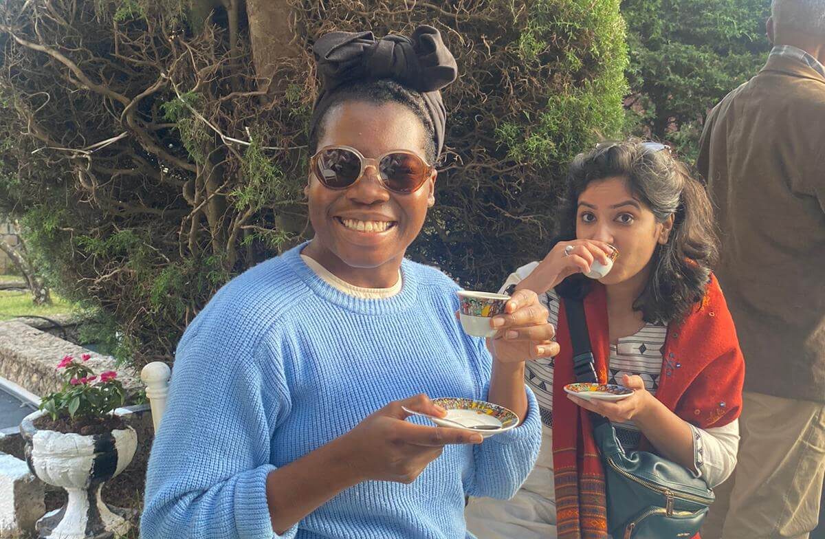 Two women sitting outdoors in a garden area enjoying coffee or tea in small cups.