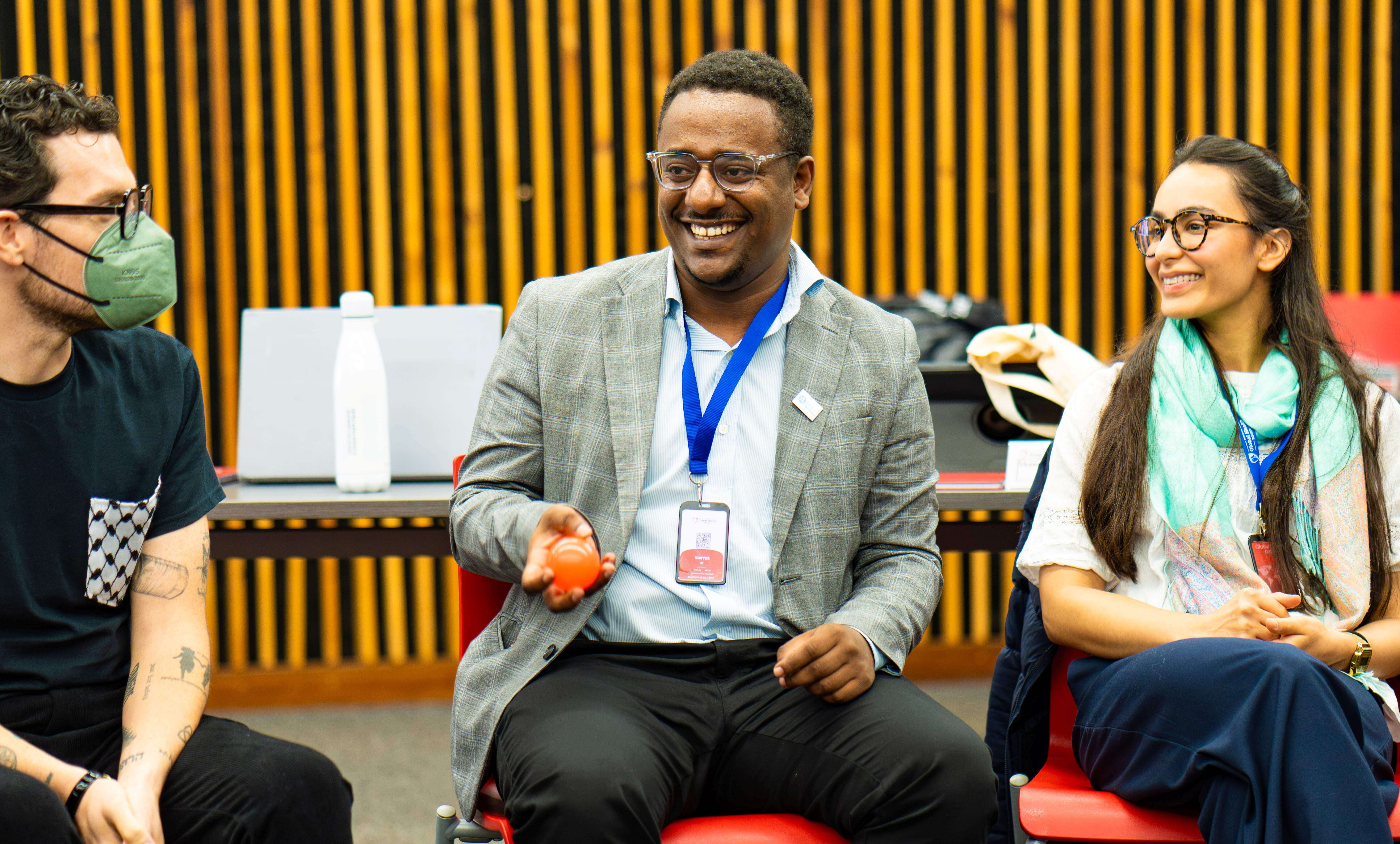 A smiling man in a grey suit jacket and blue lanyard holding a small orange ball while speaking to fellow participants seated in red chairs.