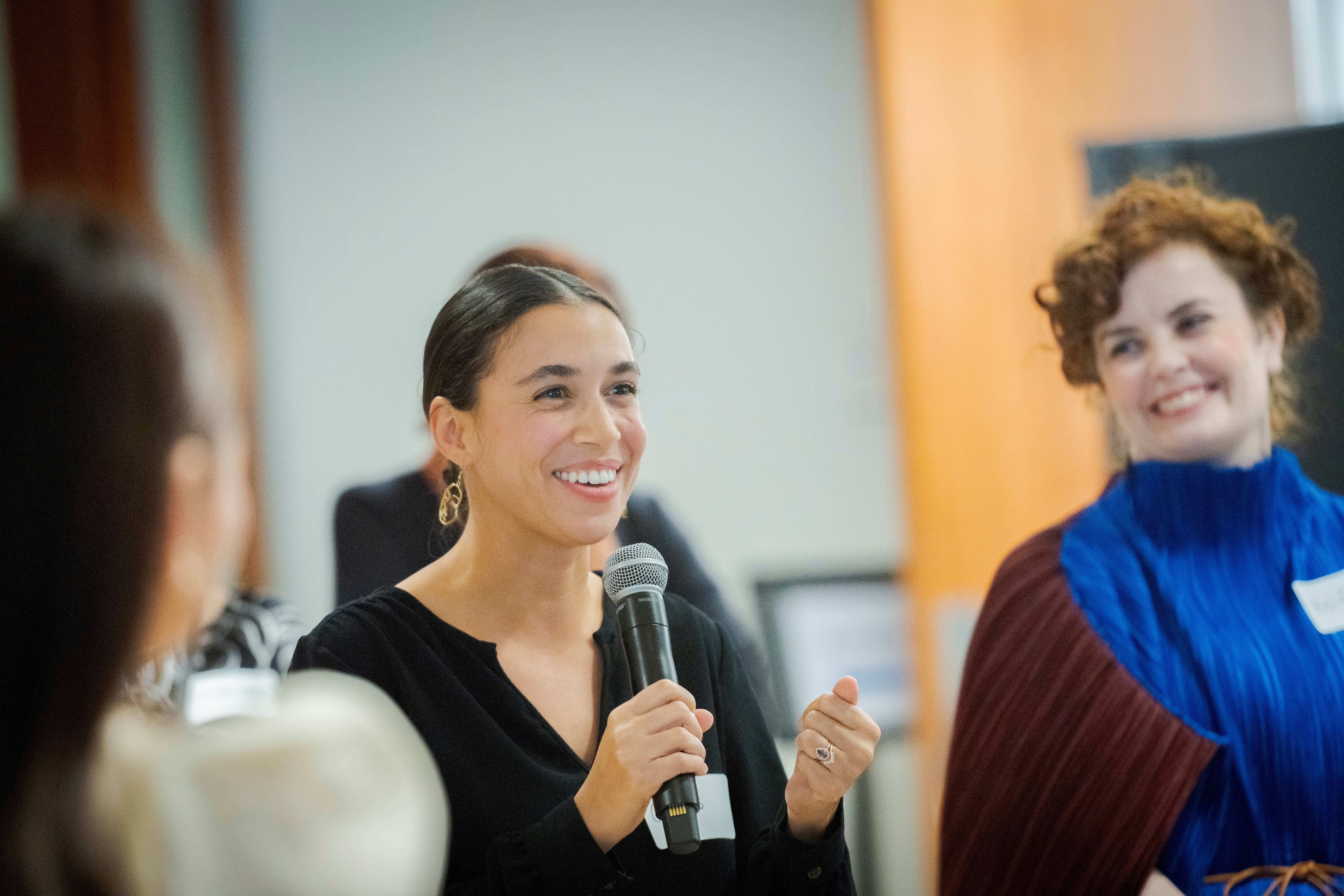 A smiling woman in black speaks into a microphone alongside an attendee in blue and red.